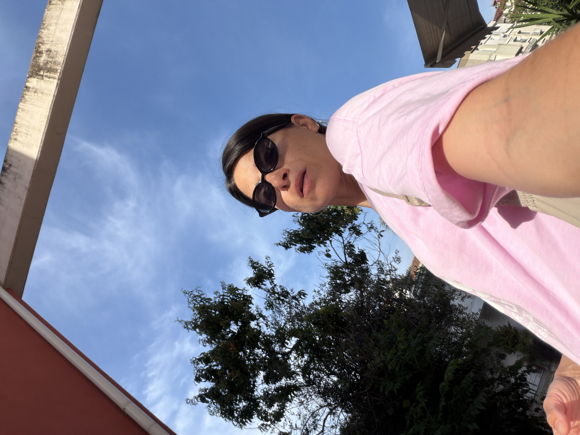 A low-angle photograph captures a woman wearing sunglasses and a pink shirt, positioned against a backdrop of a bright blue sky with wispy clouds. The scene includes a building structure in the upper left, partially obscuring the sky, and a tree with dark green foliage that fills the lower half of the frame. In the background, blurred buildings are visible. The picture appears to have been taken outdoors in Budva, Montenegro.
