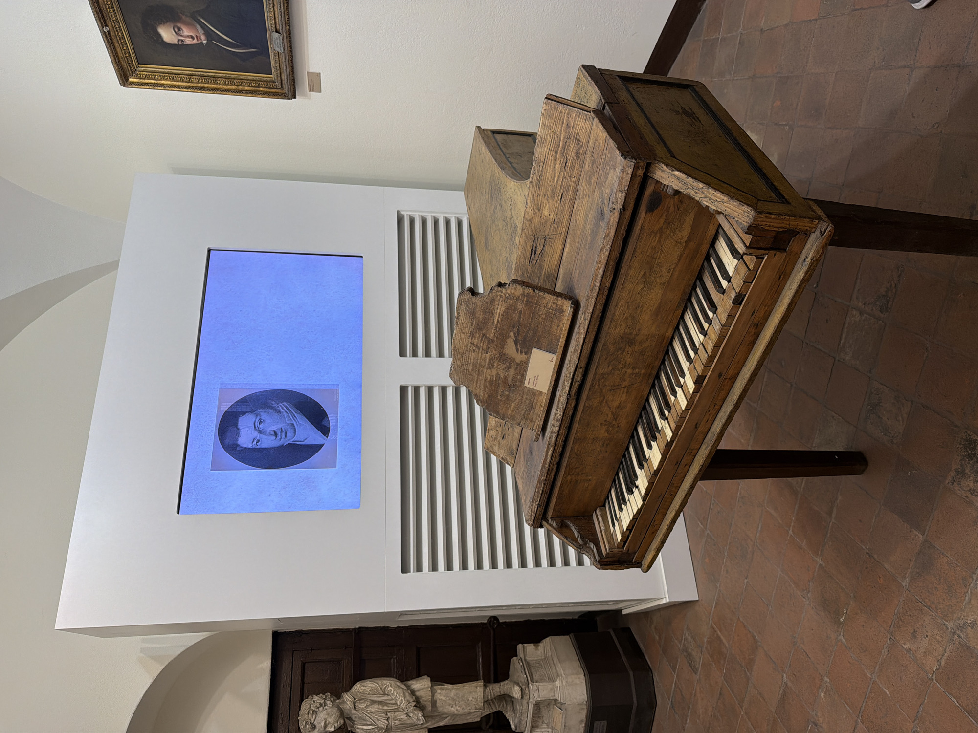 The scene is an interior shot of what appears to be a historical or cultural institution, possibly a museum, located in Catania, Italy. The floor is covered with square terracotta tiles.
The focal point is an antique, wooden piano, positioned slightly off-center. Its keys are visible, and its aged appearance suggests it is a historical artifact.
Behind the piano is a white display panel featuring a screen on which a portrait of a man is visible, presented in a blue circle. A series of vertical white slats are behind the panel on the wall.
In the background, hanging on the wall, is a framed portrait of a man.
Below the display panel, a stone sculpture of a reclining figure is visible.