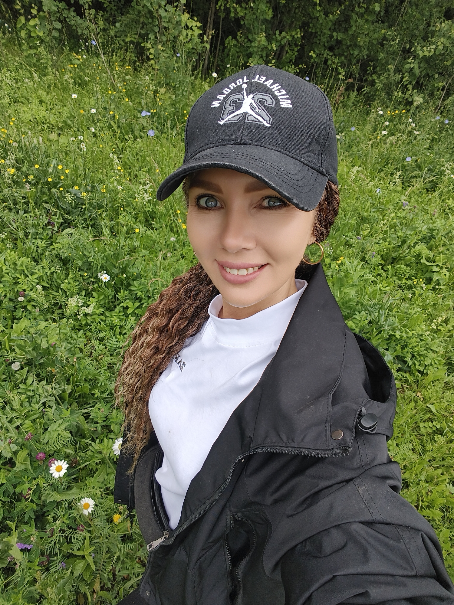 A woman with long, curly brown hair and blue eyes is taking a selfie outdoors. She is wearing a black baseball cap with white text and a logo, a white turtleneck shirt, and a black jacket. The background is a lush green field filled with wildflowers, with trees visible in the distance. The sunlight suggests it is daytime, and the greenery indicates a warm weather season. The woman is smiling at the camera, appearing happy and relaxed. The visible text on her cap reads "MICHAEL JORDAN 23". The setting appears to be a natural, possibly rural, environment in Noviy, Russia.