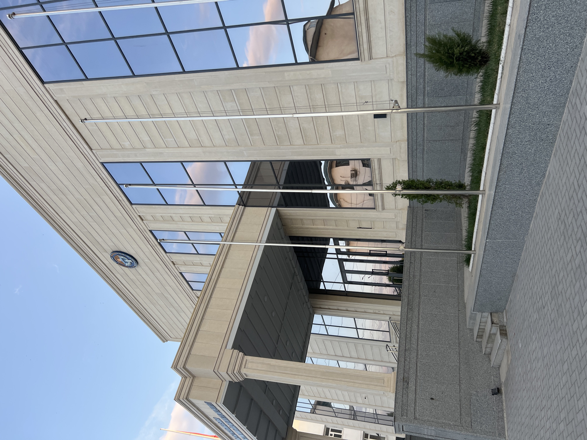 This is a full-frame, eye-level, outdoor shot of a modern, beige stone building with large, reflective glass windows in Osh City, Kyrgyzstan. The building has a grand entrance with large pillars and a covered walkway. The sky above is clear with some faint clouds and is a light blue color. The building is adorned with a circular emblem on its facade. In front of the building, there are flagpoles and small, well-maintained evergreen shrubs. A paved walkway leads to the building's entrance, with steps and a gray stone wall. The reflections in the windows show the sky and surrounding structures. No people are visible in the image.