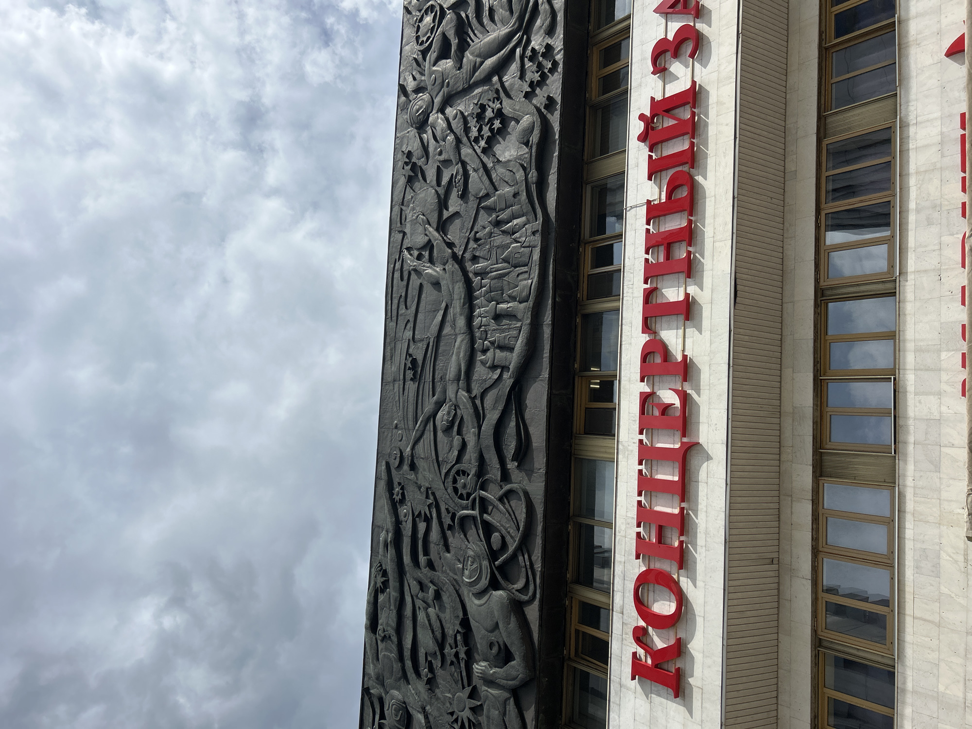 An exterior, low-angle, medium shot captures the facade of a building under a cloudy sky. The building features a large, dark, bas-relief sculpture depicting figures and celestial bodies across its upper section. Below the sculpture, bold red lettering spells out "КОНЦЕРТНЫЙ ЗАЛ" (Concert Hall) on a white marble-tiled facade. Further down, a row of rectangular windows with tan frames is visible. The overall lighting suggests an overcast day.