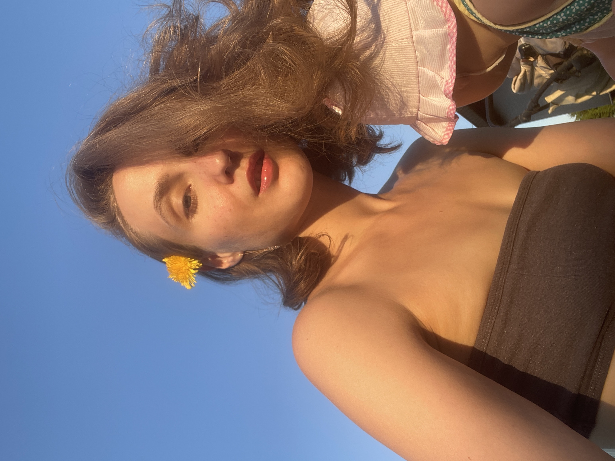A woman with light skin and brown hair is oriented vertically in the frame against a clear blue sky. A yellow flower is tucked behind her left ear. She is wearing a dark bandeau top. Another person, partially visible, is wearing a pink and white plaid bucket hat and a green top with white polka dots. The photograph's location cannot be determined from the image, and thus, the inclusion of Подшивалово, Russia, cannot be verified.