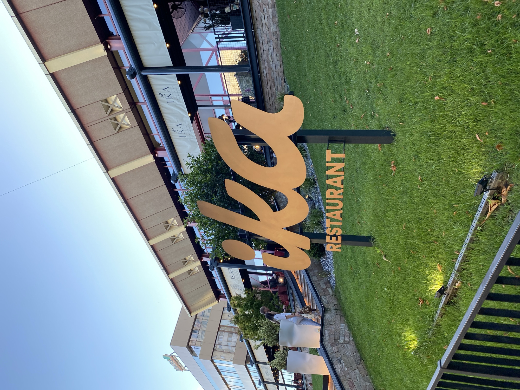 An exterior view of "IKA RESTAURANT" in Chișinău, Moldova, captured during daylight hours with clear skies. The restaurant features a prominent golden sign with the stylized letters "IKA" in large script, followed by "RESTAURANT" in block letters. The building has a modern architectural style with a sloped, segmented roof and large windows. Two awnings with the "IKA" logo are visible above the windows. In the foreground, a grassy lawn slopes upwards towards the restaurant, with scattered autumn leaves on the grass. A low black metal fence runs along the bottom edge of the frame. A couple is seen walking up a stone staircase on the left side of the image, towards the restaurant entrance. In the background, a taller, modern office building with a sign that reads "DONARIS GROUP" is visible. The scene is illuminated by natural sunlight, creating shadows on the grass.