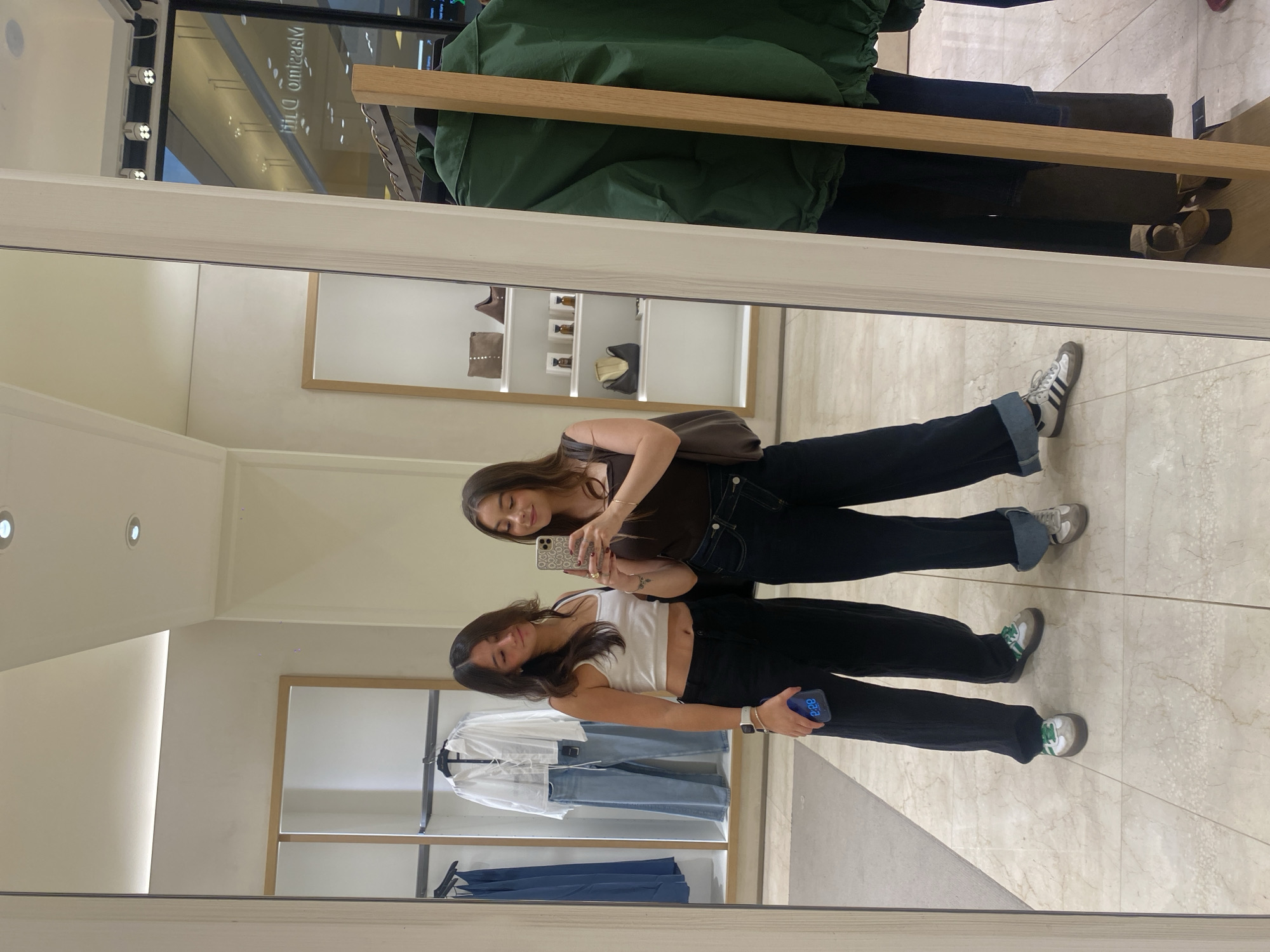 Two young women are standing side-by-side in what appears to be a clothing store, as seen in a mirror reflection. The woman on the left wears a white tank top and black pants, holding a phone in her hand, while the other woman wears a brown tank top and black pants. Clothing racks with folded clothes and hanging shirts are visible in the background. The floor has a tiled pattern. Unknown city, Unknown location.