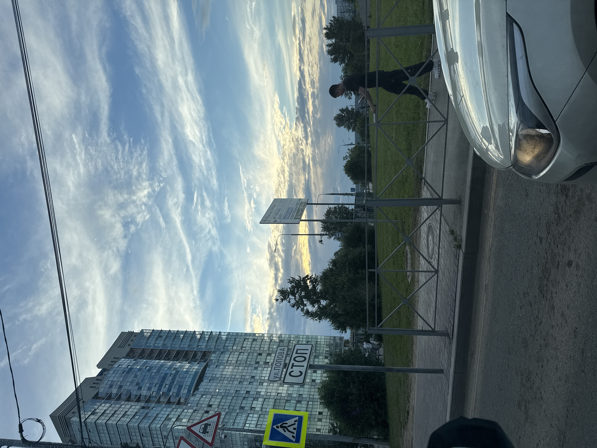 The image depicts a street scene in Perm, Russia, during what appears to be late afternoon or early evening, given the warm glow of the sky. The sky is a prominent feature, filled with wispy, elongated clouds set against a bright blue backdrop, suggesting fair weather with some atmospheric movement.
In the foreground, a modern glass-fronted building dominates the left side of the frame, reflecting the sky and its surroundings. To the right, a portion of a white car is visible, its headlight illuminated. A metal fence runs along the sidewalk, separating the road from a grassy area with trees.
Several traffic signs are present: a yellow diamond-shaped sign with a pedestrian symbol, a triangular sign with a pictogram of a basket, and a rectangular sign with text. The rectangular sign has two parts. The upper part contains an arrow pointing to the left and Cyrillic text, and the lower part has the word "СТОП" (STOP) in bold white letters on a black background, indicating a stop sign. Another sign above it is partially visible, with Cyrillic text.
A young person, likely a male judging by his attire, is standing near the fence, appearing to be interacting with something white, possibly a piece of paper. Their posture suggests a casual or contemplative stance.
The scene is urban, with a mix of modern architecture, road infrastructure, and greenery. The overall mood is calm, possibly hinting at the end of a day's activities. The presence of traffic signs and the car suggest this is a public street or area.