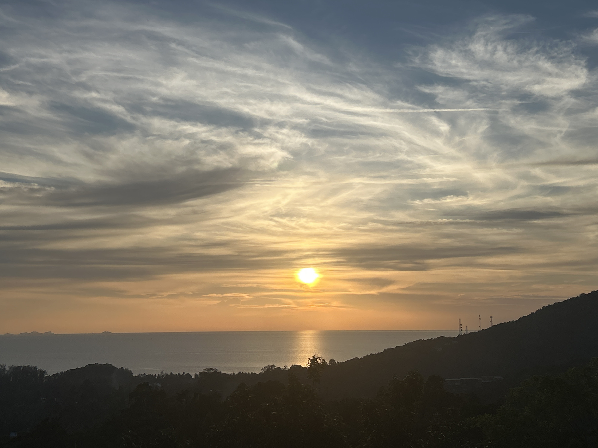 The image shows a sunset over the sea in ตำบลเกาะพะงัน, Thailand. The sun is centered in the frame, reflecting on the water. The sky is filled with streaky clouds, ranging from white to gray. The foreground features silhouetted trees and a hill with antennas. The sea horizon displays indistinct islands.