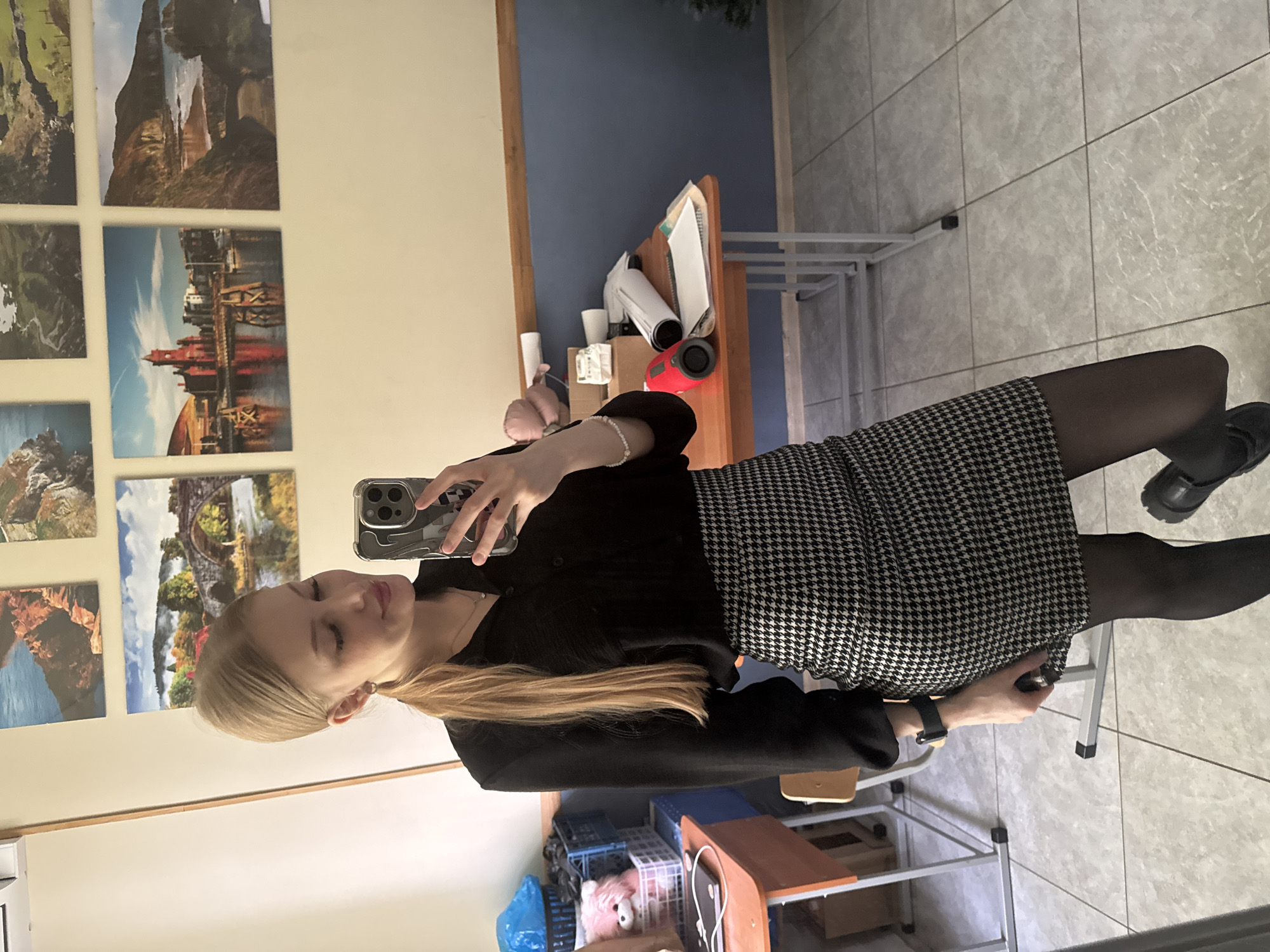 A woman is taking a selfie in a room. She has long blonde hair and is wearing a black shirt, a black and white patterned skirt, black tights, and black shoes. The background includes a wall with several landscape pictures, a desk, and some storage shelves. The location cannot be determined as the picture does not include any identifying information that indicates this is Perm, Russia.