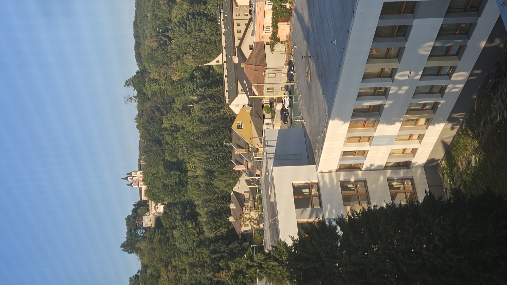 The image captures a sunny daytime scene in Prešov, Slovakia, overlooking a town nestled at the base of a forested hill. In the distance, atop the hill, a prominent building with a red roof and two pointed spires, possibly a church or a monastery, is visible against a clear blue sky. The midground features a cluster of residential buildings with various rooflines and colors, interspersed with trees. In the foreground, a modern, multi-story white building with many windows dominates the lower portion of the frame, casting long shadows. A large, dark green conifer tree is situated to the left of this building. The overall impression is one of a peaceful urban landscape merging with nature.