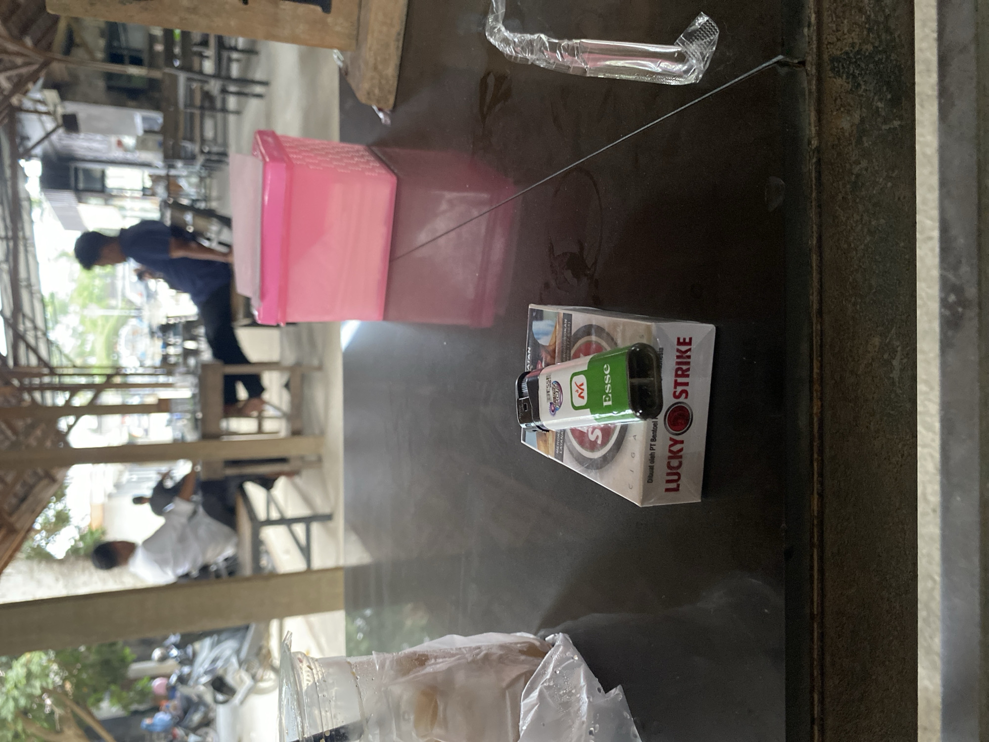 The image captures a casual indoor or semi-outdoor setting, likely a cafe or eatery, in Gampongaceh, Indonesia. The perspective is from a table, looking towards the back of the establishment.
In the immediate foreground, resting on a dark table surface, are several objects: a clear plastic cup containing a beverage and wrapped in plastic, a green and white cigarette lighter, and a pack of Lucky Strike cigarettes. A clear plastic straw or tube is also visible near the edge of the table. The cigarette pack prominently displays "LUCKY STRIKE," "Dibuat oleh PT Bentoel," and elements like "JESSE" and "Esse." The lighter also features the "Esse" brand logo.
Further back on the table, there is a pink plastic container. The background is blurred, showing several people and more tables and chairs. One individual in a blue shirt and dark pants is seen standing, while another in a white shirt is seated. The architecture includes wooden beams, suggesting an open or traditional structure. Beyond the seating area, hints of an outdoor space with parked motorbikes are visible. The scene portrays a daytime environment where people are gathered, possibly for dining or socializing, with smoking materials present on the table.