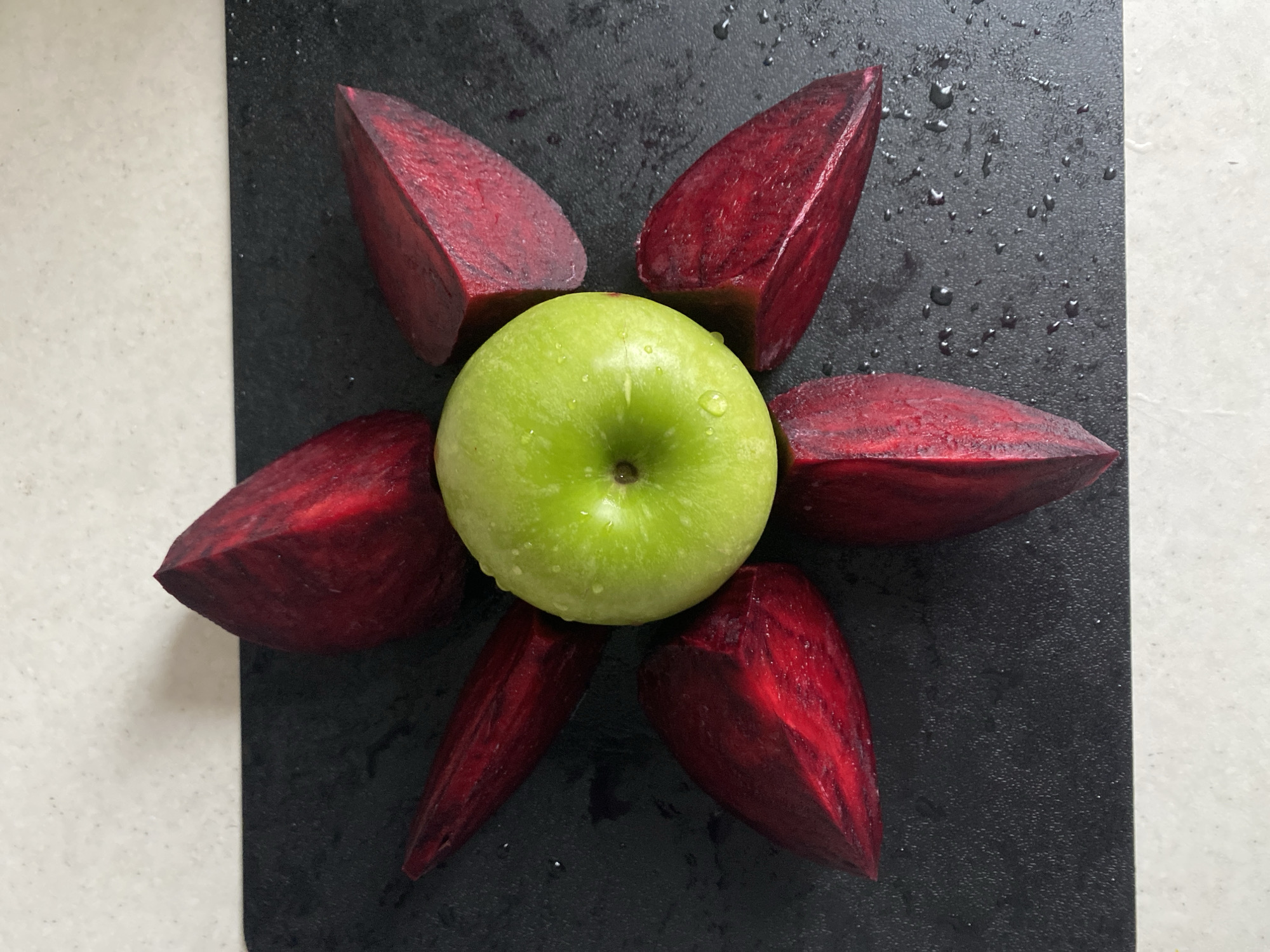 A top-down, medium shot reveals a green apple positioned in the center, surrounded by five wedge-shaped segments of red beets on a black cutting board. The scene is likely indoors, based on the consistent lighting. The cutting board is placed atop a countertop. No people are visible. There is evidence of moisture, with water droplets present on both the apple and the cutting board. The beets and apple are the primary subjects. The location is not discernible from the visual information.