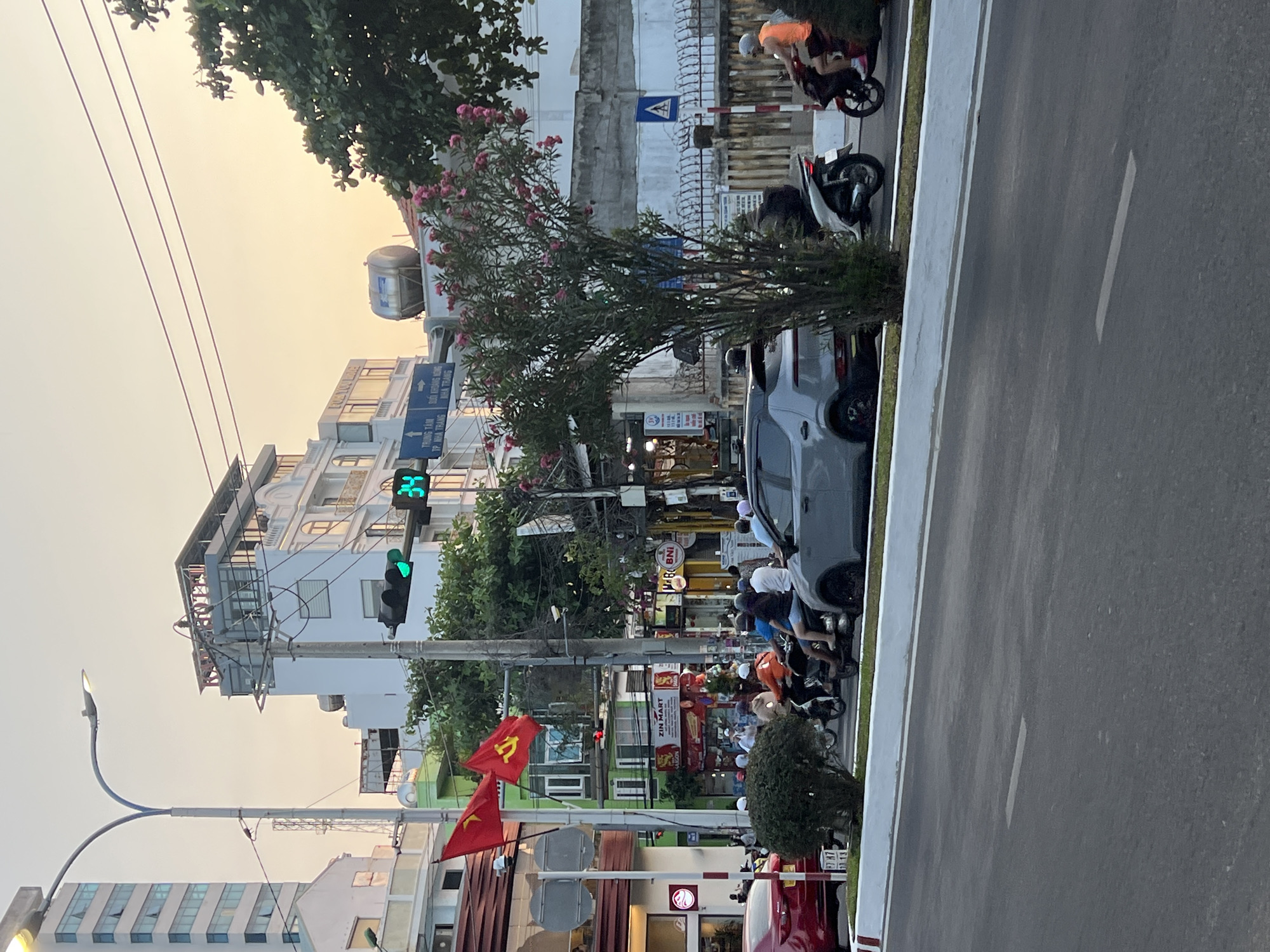 The image depicts a street scene in Bắc Nha Trang, Vietnam. A grey car is parked on the side of the road. Several motorcycles with people are stopped at a green traffic light. A street lamp is visible on the left. Buildings line the street, and red flags are displayed. Street signs provide directions to "TRUNG TAM TP. NHA TRANG" and "SUOI KHOANG NONG NHA TRANG". A convenience store called "ZIN MART" is visible.