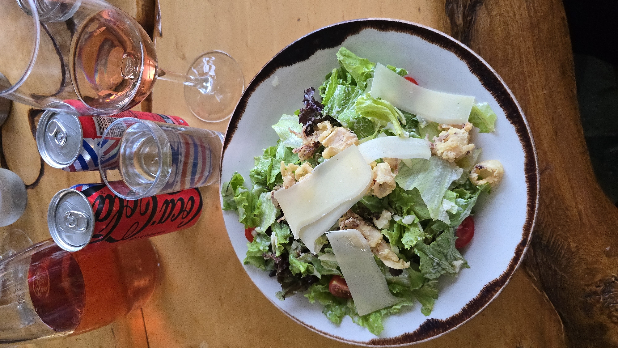 A close-up, eye-level shot captures a meal setting on a wooden table. In the foreground, a white bowl with a dark rim holds a fresh salad composed of various leafy greens, cherry tomatoes, crumbles of fried ingredients, and thin shavings of white cheese. To the right of the salad, a stemmed wine glass contains a rose-colored liquid. Behind the salad, a clear glass is filled with water, and two soda cans, one red with white lettering and the other with a red, white, and blue design, stand upright. A glass pitcher filled with a reddish-pink liquid is visible in the upper left corner. The background is blurred, hinting at a casual dining environment. The image does not contain any information about the specific location in Krasnovo, Bulgaria.
