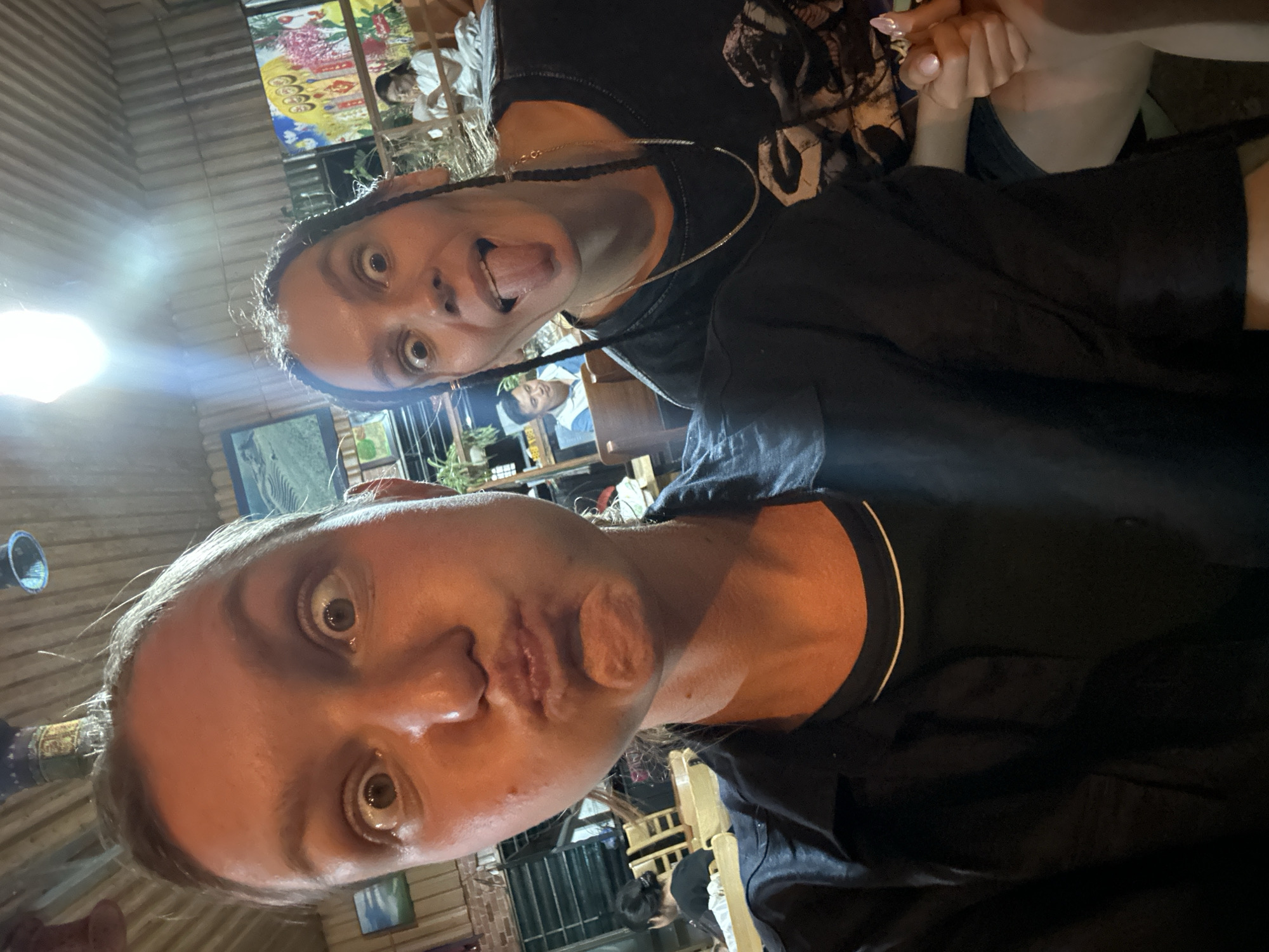 The image is a playful selfie featuring two individuals in a dimly lit indoor or semi-outdoor setting, likely a restaurant or cafe in Mù Cang Chải, Vietnam.
In the foreground, a person with light hair has wide, surprised eyes and pursed lips. They wear a dark shirt with a light trim around the collar. Behind them, another person with dark braided hair sticks their tongue out, also with wide, mischievous eyes. This individual wears a dark t-shirt with a subtle pattern and a thin chain necklace. Their hands are clasped with another person's hand, suggesting a close or friendly interaction, and one hand shows light pink nail polish.
The background shows other patrons seated at tables, largely blurred. The setting has a rustic feel, featuring wooden ceiling panels and beams. Colorful artwork or posters, some with floral patterns and stylized faces, adorn the walls. Potted plants are visible throughout the space. A bright light source in the upper left creates a strong glare, indicating that the photo was taken at night or in the evening. The overall mood is casual and lively. No legible text is clearly visible in the image.