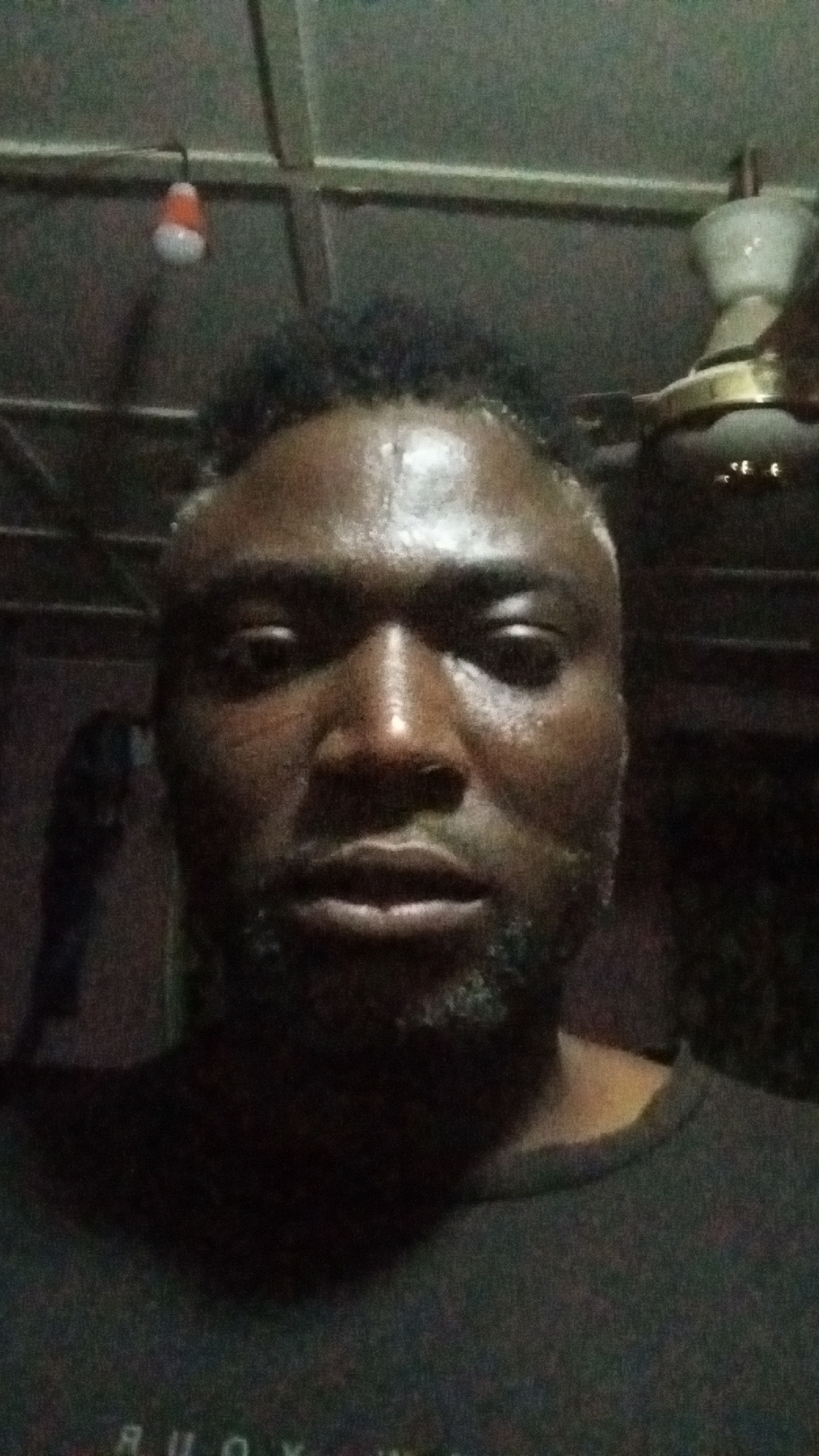 The image is a close-up selfie of a man with dark skin and short, dark hair, some visible beard, and moist skin. He is wearing a dark-colored t-shirt. The background is dark, but includes a ceiling with a light fixture and a ceiling fan. The location, Sabon Tasha, Nigeria, is provided, but cannot be confirmed from visual information within the image.