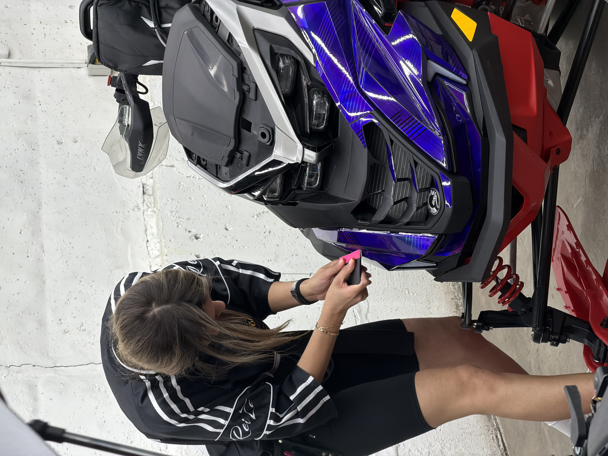 A woman is applying a graphic wrap to the front of a snowmobile. The snowmobile is predominantly purple, white, silver, black, and red. The woman is wearing a black top with white stripes on the sleeves, black shorts, and a watch on her left wrist. She is holding a pink squeegee and applying it to the purple wrap on the snowmobile. The background is a white cinder block wall. The location is most likely in an indoor workspace, such as a garage or shop. The image does not provide sufficient information to determine whether the location is in Krasnoyarsk, Russia.