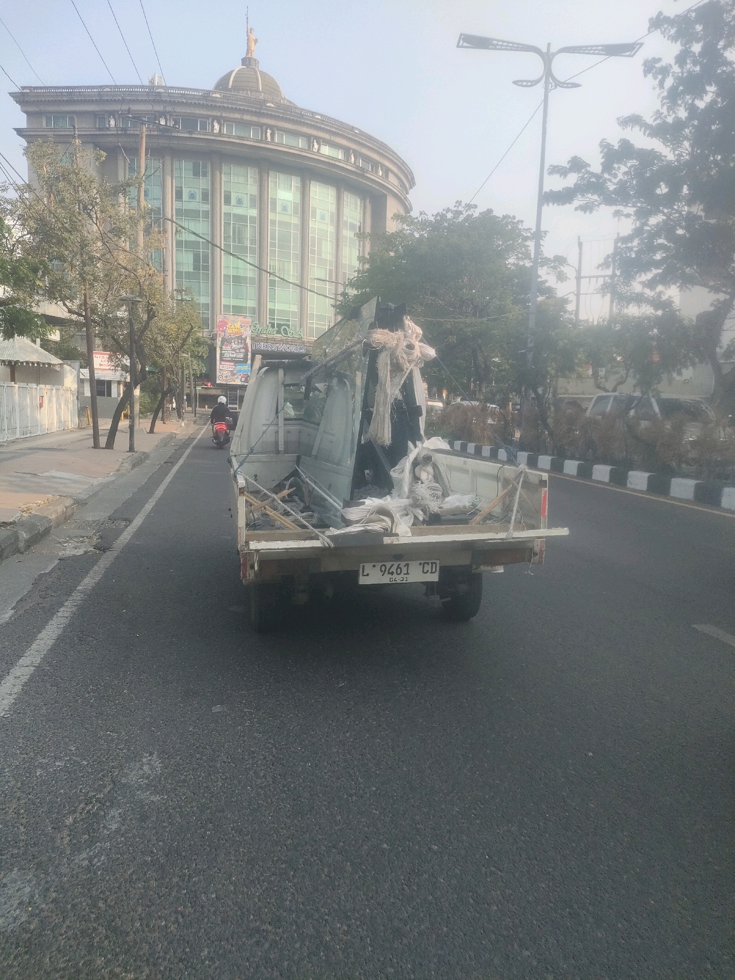 In Surabaya, Indonesia, a slightly hazy, overcast day is depicted. The foreground is dominated by the rear of a white pickup truck, laden with debris that includes white plastic sheeting and what appears to be construction materials or packing waste. The truck's license plate reads "L 9461 CD". The truck is on a paved road with a white dashed line marking the lane.
To the left of the road, a sidewalk is visible with a low curb. A white fence and a building with a white exterior are seen behind the sidewalk. A lone motorcyclist wearing a white helmet and riding a red scooter is proceeding away from the camera down the same road, in the same direction as the pickup truck.
Further down the road, a large, curved, multi-story building stands out. It has a distinctive dome topped with a statue resembling the Statue of Liberty. The building's facade is predominantly glass windows framed by white architectural elements. Trees with sparse foliage line the area in front of this building, and several power lines are strung across the upper portion of the image. To the right of the road, trees and other vegetation are visible, with some indication of other vehicles in the distance obscured by the haze. A modern street lamp with a distinctive arching design is positioned on the right side of the road. The overall scene suggests an urban environment undergoing some activity, possibly related to construction or waste removal.
