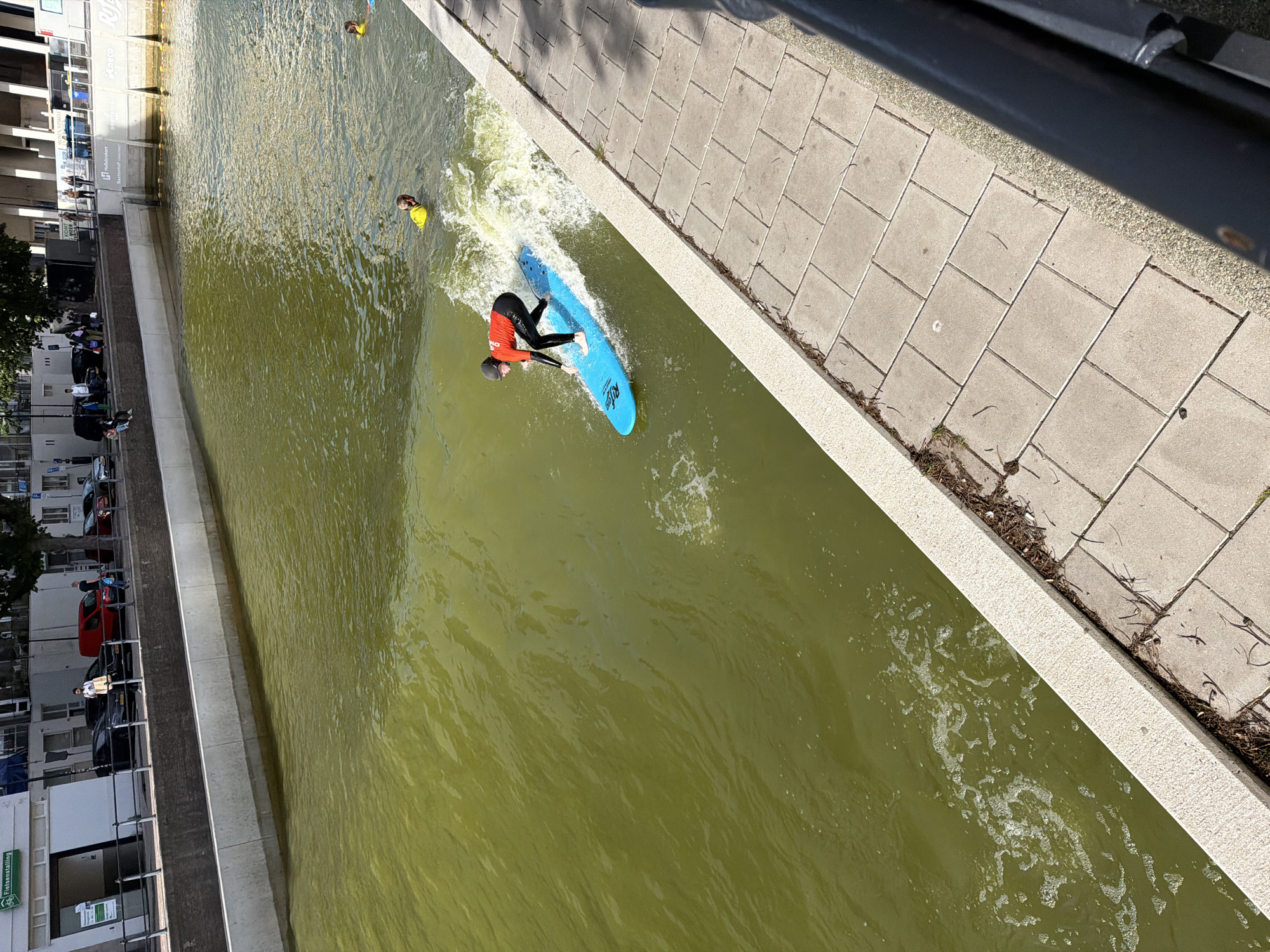 In Rotterdam, Netherlands, a person in a red shirt and black wetsuit surfs on a blue surfboard in a canal. The canal has murky green water and is surrounded by a concrete embankment and a tiled walkway. Another person in a yellow vest is visible further in the water. The background shows buildings and trees, suggesting an urban environment.