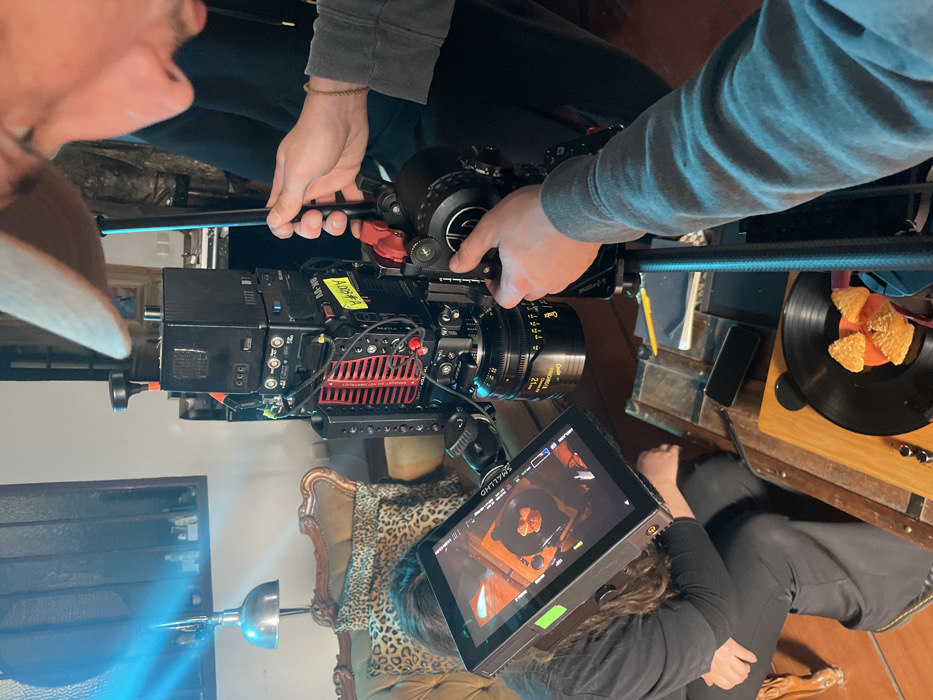 The image depicts an indoor film production scene. A professional digital cinema camera, featuring a prominent red heatsink, multiple stacked power and transmission modules, and a Quasar Science 21mm cine lens, is centrally positioned on a tripod or stand. A person, partially visible on the right, wearing a beige cap and a grey long-sleeved shirt, is operating the camera, with both hands adjusting controls on the camera's baseplate and follow focus unit. To the left, another individual with long dark hair, dressed in dark attire, is seated or crouched, intently observing a SmallHD 702 Touch monitor which displays the camera's live feed. The monitor's display shows a close-up shot of a vintage turntable with a black vinyl record, upon which several pieces of fried food (resembling chicken nuggets) are arranged. The camera is pointed downwards, capturing this specific arrangement on the turntable. In the background, a vintage-style sofa with ornate wooden framing and leopard print cushions is visible. A wooden trunk or chest serves as a surface, holding a smartphone and a yellow pencil. A silver dome lamp is present in the background, and a distinct blue light beam diagonally illuminates the upper left portion of the frame, indicating artificial lighting for the set. A third person in dark clothing is partially visible behind the main camera operator. The specific location within Paris, France, is not discernible from the visual information presented in the image.