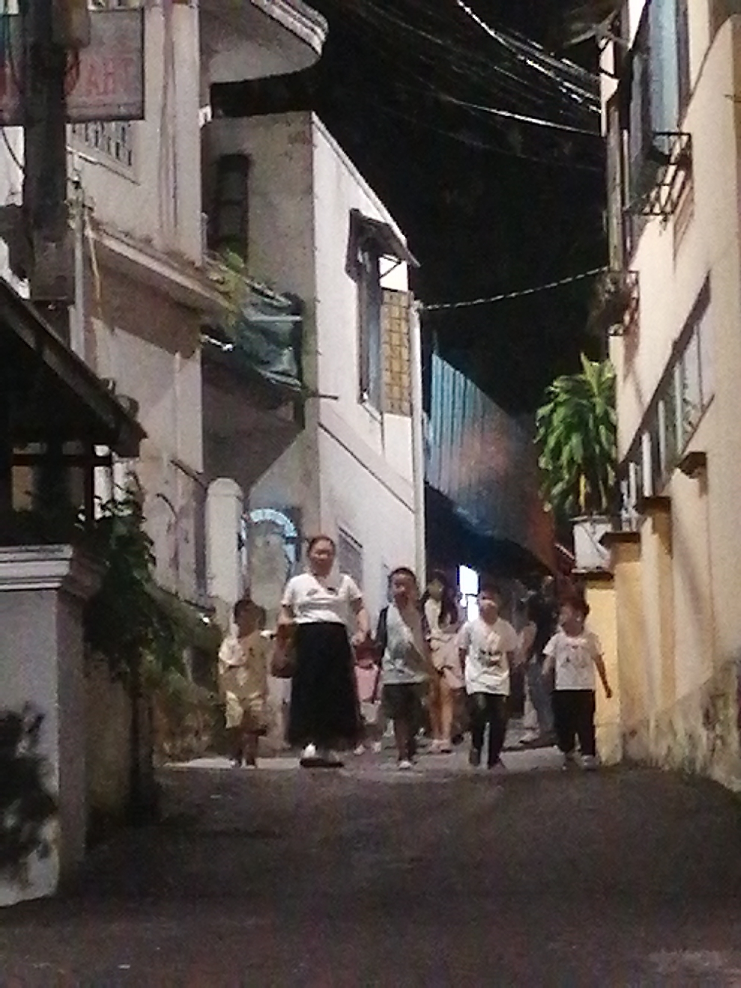 The image depicts a narrow street scene at night in Phường Xuân Hương, Đà Lạt, Vietnam. Several people are walking along the street. In the foreground, a woman wearing a white shirt and black skirt is visible, her body is partially obscured by the foreground. To her left, a child in a yellow outfit is walking. Further down the street, several other individuals, including children wearing white t-shirts, are walking in the same direction. The street is lined with buildings on both sides. The building on the left appears to be a light-colored multi-story structure with visible windows and some architectural details. The building on the right is also light-colored and appears to be an apartment building or commercial establishment. Overhead, electrical wires are strung across the street. The overall lighting suggests it is nighttime, with artificial illumination from within the buildings and possibly streetlights. The ground is paved.