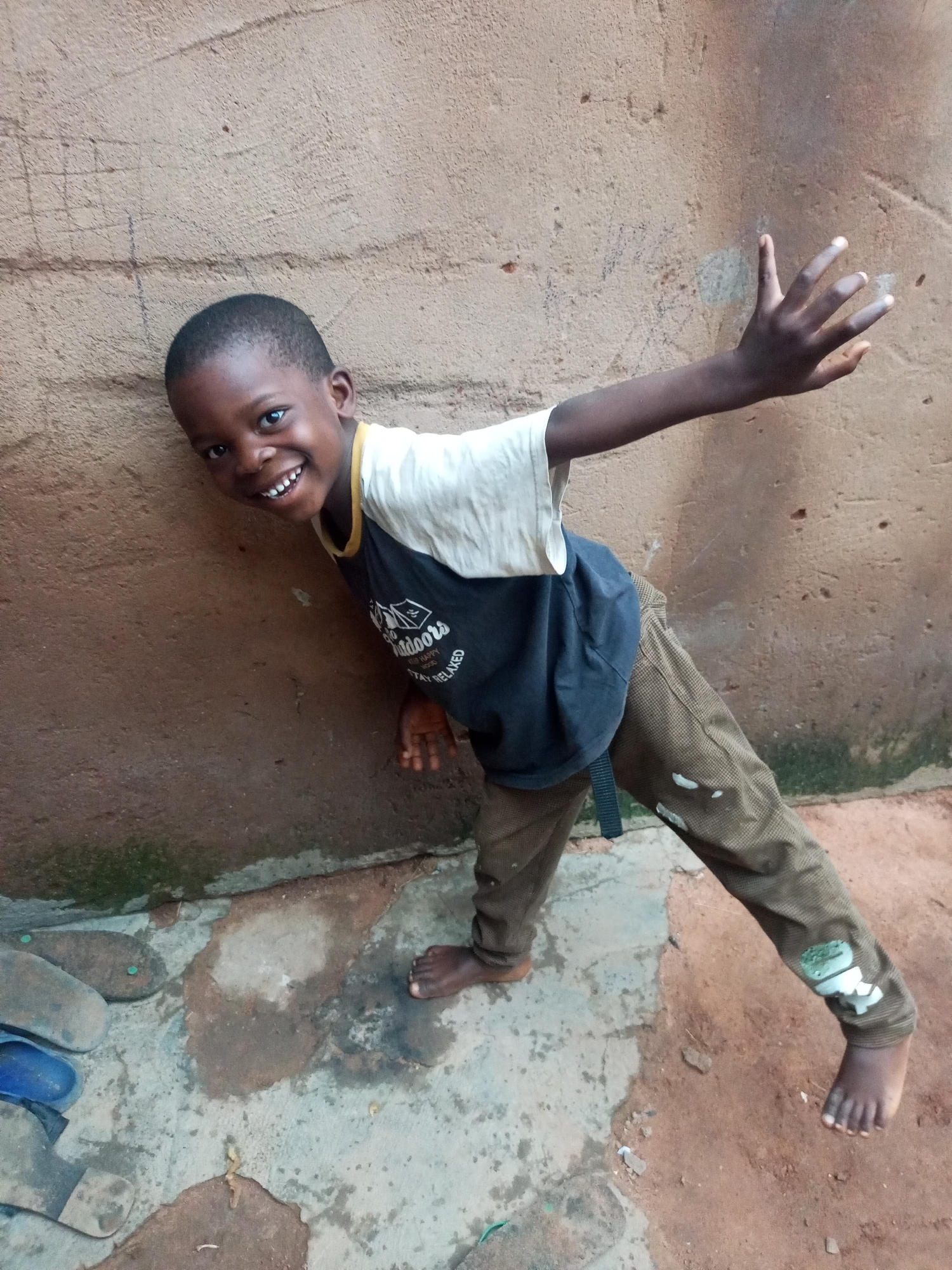 In Tumfure, Nigeria, a young boy with dark skin and short hair is captured in a candid outdoor photograph. He is leaning against a rough, textured, light brown wall, his body angled towards the viewer. The boy wears a dark t-shirt with a light-colored collar and a white shirt underneath. His pants are a muted brown with a subtle pattern and some white marks. His right arm is extended upwards and outwards, with his hand open, fingers spread, as if reaching or waving. His left arm is bent and partially visible. The boy's face is turned towards the camera, and he is smiling broadly, revealing his teeth. His eyes are open and looking directly at the lens.
The ground in front of him consists of a mix of concrete and reddish-brown earth. There are some discarded flip-flops and a blue sandal visible on the left side of the frame. Patches of green moss or algae are present on the wall and the ground, indicating a damp environment. The lighting suggests it is daytime, with a slight shadow cast on the wall. The overall impression is one of a playful and energetic child in a simple, everyday setting.
