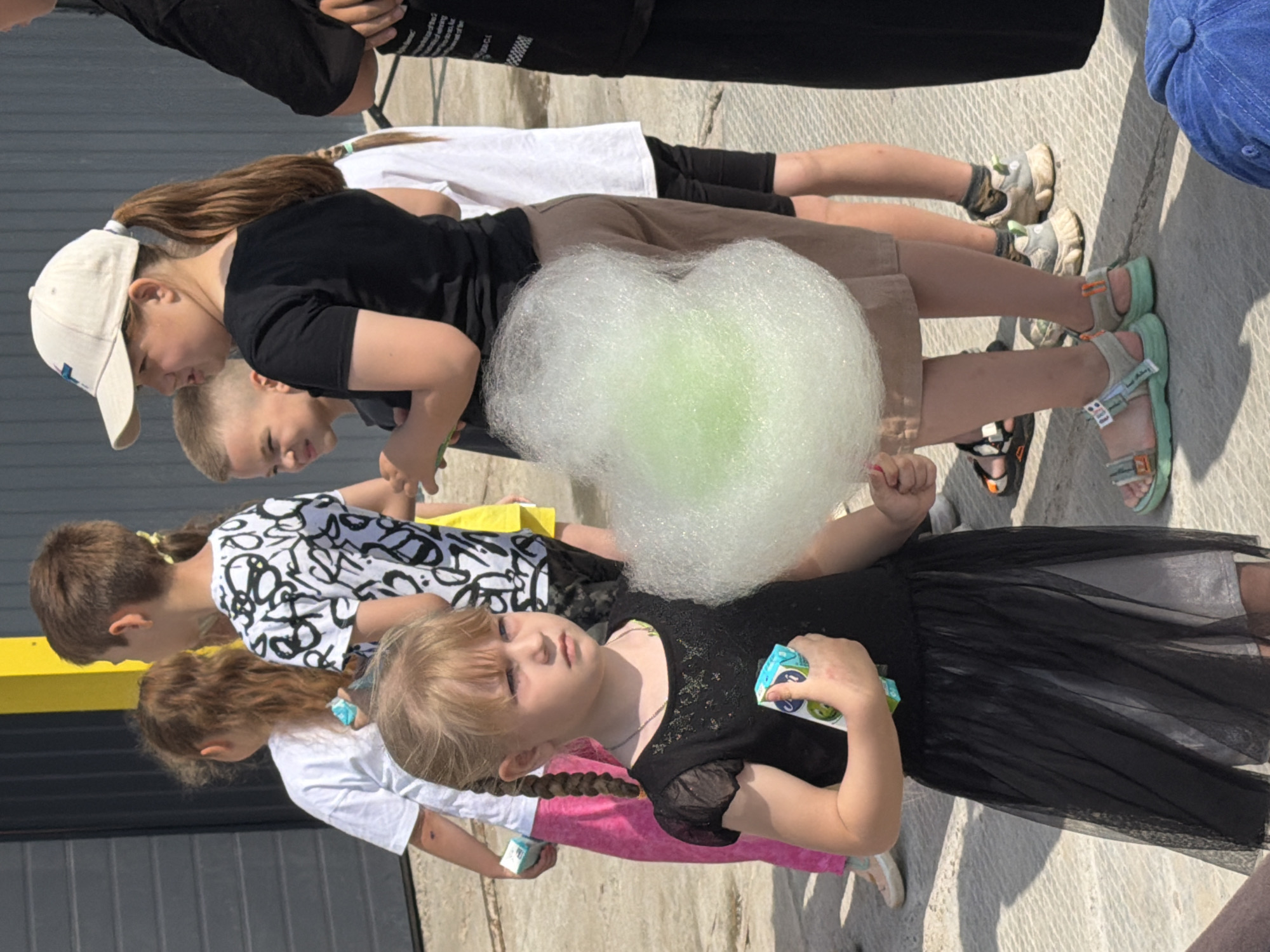 A group of children and adults stand outside on a sunny day, with a light gray concrete ground and a dark gray metal fence in the background. A yellow pole is visible to the left of the frame. The foreground is dominated by a young girl with blonde hair in braids, wearing a black dress and holding a light green cotton candy. She is also holding a small juice box. To her right, a girl with a ponytail and a white baseball cap, wearing a black t-shirt and brown shorts, is looking to the left. Behind her, a boy with brown hair is looking forward. Further back, other people are visible, including a boy in a white t-shirt with black markings and a girl in a white t-shirt and pink skirt. The overall scene appears to be a casual outdoor gathering or event. The setting is the city of Chaikovsky, Russia.