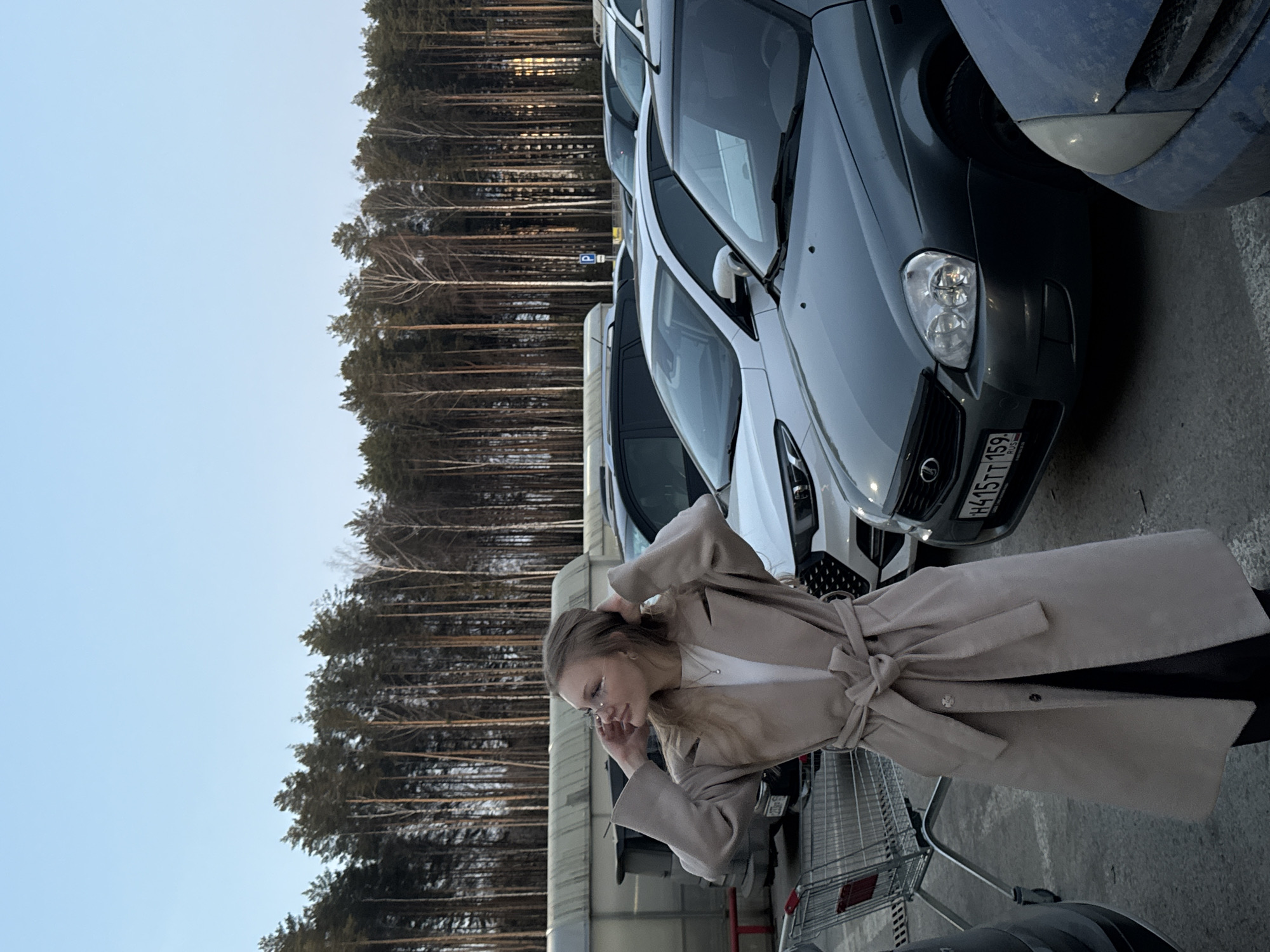 A woman in a beige trench coat stands next to a shopping cart in a parking lot in Perm, Russia. She has her hands near her head, possibly adjusting her hair or glasses. The parking lot is adjacent to a dense forest and filled with several cars. The license plate "H415TT 159 RUS" is visible on one of the vehicles. The sky is light blue.