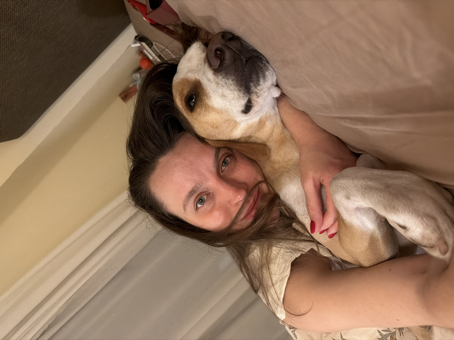 An adult human female and a dog are depicted in an intimate indoor setting, likely a bedroom. The female, with long dark hair and light eyes, directly faces the camera, her facial expression appearing neutral to slightly smiling. She is wearing a light-colored top and has red nail polish on her visible right hand. Her right arm embraces a medium-sized dog, which is primarily fawn/tan and white with brown floppy ears and a dark muzzle patch. The dog's head rests comfortably on a light brown/tan pillow or bedding, nestled against the female, with its eyes partially closed or looking downwards. Both subjects are covered by light brown/tan bedding. In the background, to the left, light-colored curtains or blinds are visible. To the right, a dark brown headboard or wall panel is present, with indistinct objects, including a red spherical item and some wiring, visible on a surface behind it. The lighting is soft and warm. The provided contextual information places this location in the city of Budva, Montenegro.