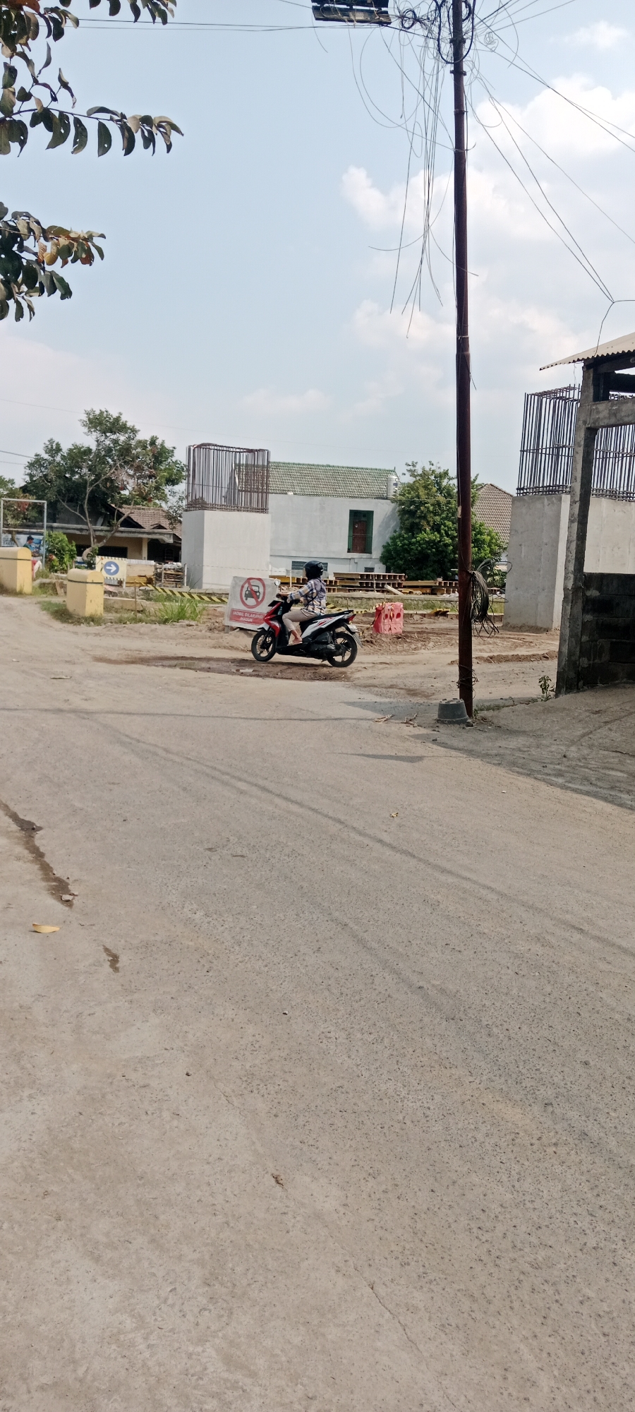 In Purwomartani, Indonesia, a person rides a scooter on a paved road. The scooter is red and white, and the rider is wearing a helmet and a patterned shirt. The scooter is in motion, as indicated by the blurred wheels. In the background, there is a construction site with concrete pillars and rebar, suggesting infrastructure development. A white building with a green roof is visible behind the construction. Several trees are scattered throughout the scene. A utility pole with numerous wires stands to the right of the frame. The sky is clear with a few scattered clouds. The road surface shows signs of wear and tear, with some cracks and dark patches indicating moisture. A sign with a red circle and a diagonal line, indicating prohibition, is visible near the construction.