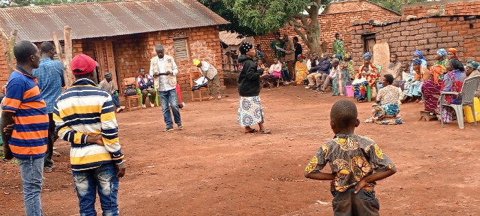A gathering of people, mostly women, is seated outdoors in a circle on chairs and mats. A woman in a black jacket and patterned skirt stands and addresses the group. Several men and young boys stand outside the circle, observing the scene. The setting appears to be a rural village, with brick buildings and a dirt ground. One man stands and appears to be taking notes with a tablet.