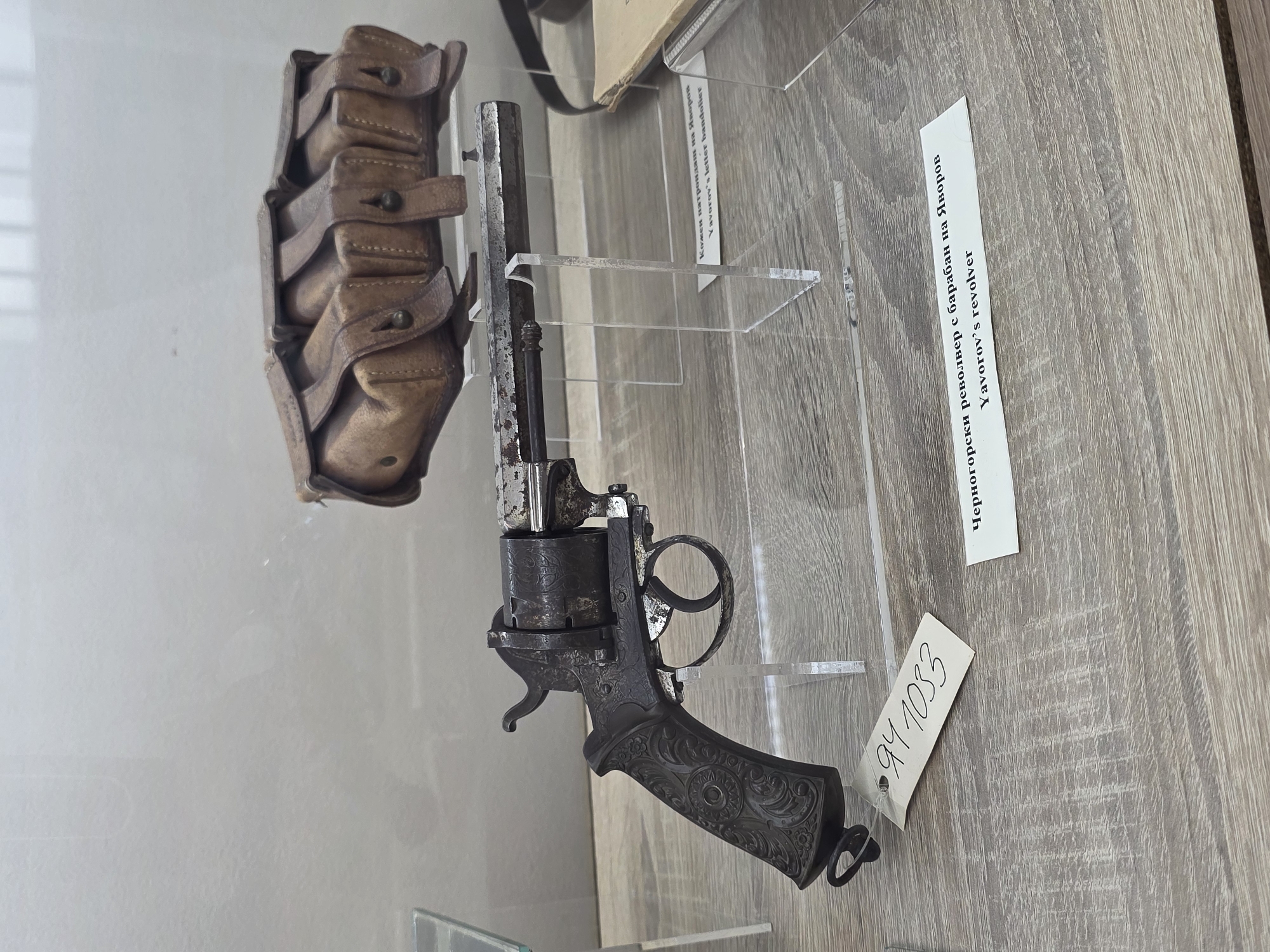 The image displays a museum exhibit featuring historical weaponry and accessories. In the foreground, a tarnished, ornate revolver with a cylinder is presented on a clear acrylic stand. A white tag with handwritten numbers "941033" is attached to the revolver's grip. Behind the revolver, a brown leather bandolier with multiple pouches is also displayed on an acrylic stand. A label in both Cyrillic and English identifies the items as "Черногорски револвер с барабан на Яворов Yavorov's revolver" and "Кожен патрондам на Яворов Yavorov's letter bandolier". The background shows a light-colored, textured wall and a wooden surface. The context provided by the labels suggests this exhibit is located in Chirpan, Bulgaria, and pertains to items associated with Yavorov.
