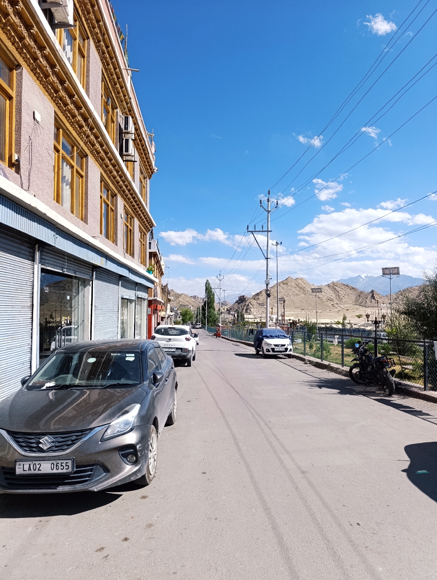 The image depicts a street scene in Leh, India, under a clear blue sky with scattered white clouds. The perspective is from street level, looking down a paved road lined with buildings and natural scenery.
On the left side of the frame, a multi-story building with a light-colored facade and ornate yellow trim runs along the street. The lower level features closed storefronts with grey roller shutters and some glass windows. Air conditioning units are visible on the upper levels.
The street itself is asphalt and appears to be a public thoroughfare. Several vehicles are present:
- In the foreground, a dark grey Suzuki sedan is parked, with its front license plate reading "LA02 0655".
- Further down the street, a white SUV is parked.
- A white car is parked on the right side of the road, partially covered.
- A black motorcycle is parked on the right side of the road, near a fence.
- In the distance, other vehicles and a few people are visible.
The right side of the street is bordered by a wire mesh fence and vegetation, beyond which are hills and mountains. The landscape is arid, with rocky terrain and sparse greenery. In the far distance, snow-capped mountains are visible under the bright sky. Power lines and utility poles traverse the scene, indicating the presence of infrastructure. The overall impression is of a somewhat quiet, sunny day in a town located in a mountainous region.
