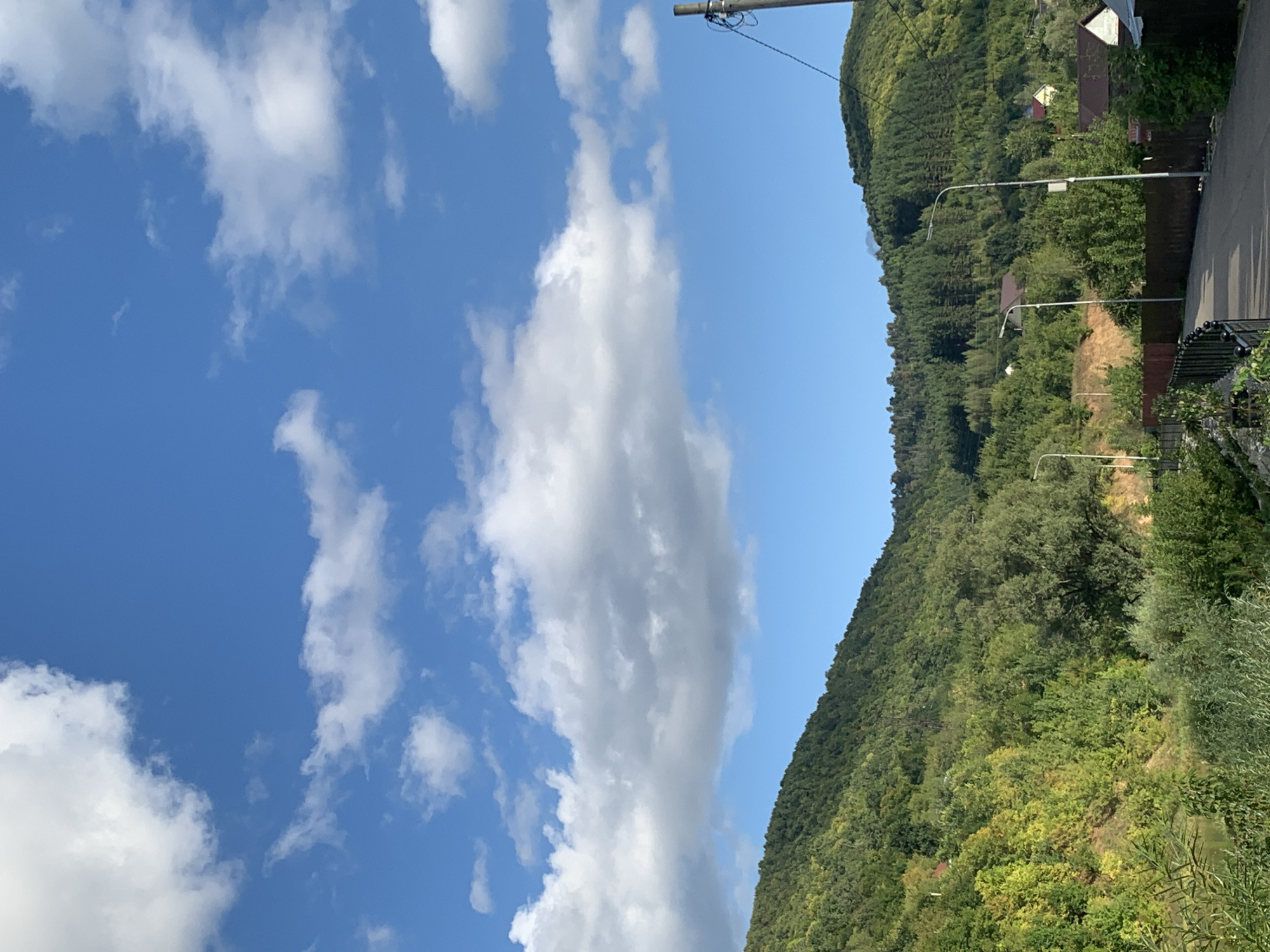 A sunlit, wide-angle shot captures a serene natural landscape with a clear blue sky and scattered white clouds above rolling green hills. The hills are densely covered with trees, exhibiting various shades of green and hints of yellow, suggesting late summer or early autumn. In the foreground, a paved road runs diagonally from the bottom right towards the center, bordered by a dark brown fence and streetlights. Some houses with red roofs are visible nestled amongst the trees on the slopes of the hills. A body of water, possibly a river, is partially visible on the left, surrounded by lush green vegetation. The image appears to be taken in a rural or suburban area, possibly on the outskirts of Asha, Russia, given the characteristic hilly terrain and vegetation.