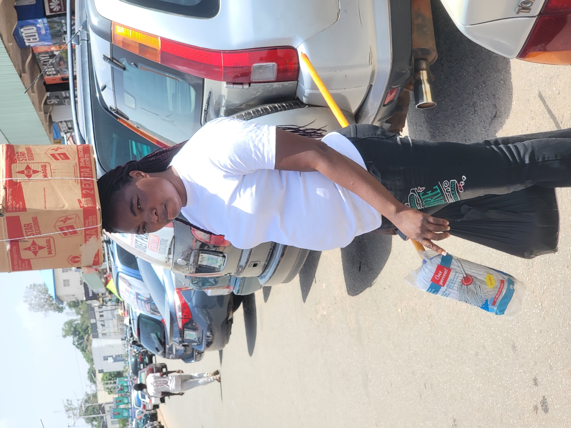 A young woman, appearing to be a street vendor or market seller, stands on a paved street in what looks like an urban setting, likely in Jos, Nigeria. She is wearing a white t-shirt and dark pants with a stylized "RE JEANS" logo. Her hair is braided.
She skillfully balances a large cardboard box on her head, secured with a white strap. The box is prominently branded with "STAR SEASONING" and "MAGGI" logos, and text indicates "20g CHICKEN SACHETS" and "2 EXTRA SACHETS." In her right hand, she holds a black plastic bag and a packaged mop. The mop's packaging shows the brand "One percent" and the phrase "LA MEJOR CALIDAD."
Behind her, a silver SUV (possibly a Honda CR-V) is parked, and part of a "TOYOTA" logo is visible on another vehicle. A yellow stick or rod is propped against the rear bumper of the silver SUV. Numerous other cars are parked along the street, suggesting a busy commercial area. In the distance, other people can be seen walking, and buildings line the street.
The scene is brightly lit by natural sunlight, indicating it is daytime with clear weather. The woman looks towards the camera with a neutral expression. The way she carries the goods on her head is a common practice in the region, adding to the local context. A partial sign with "LIKET P" is visible in the background.