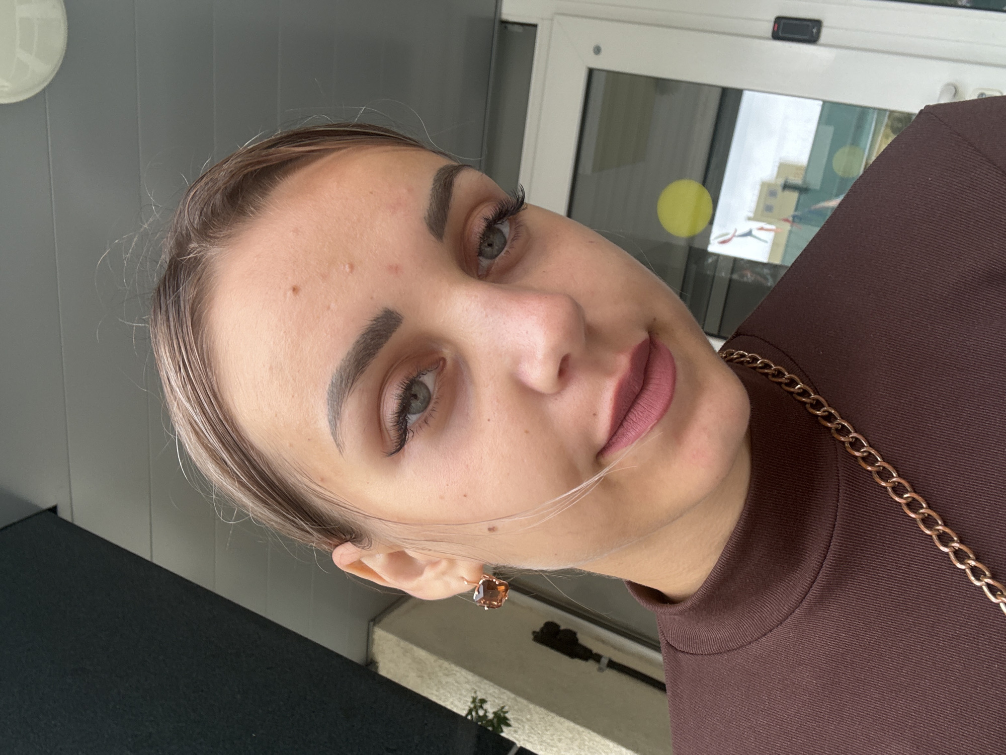 A close-up, medium-angle selfie captures a young woman outdoors, with a shallow depth of field. She is facing the camera and appears to be wearing makeup, including mascara and lipstick. She has a small mole on her forehead and some blemishes on her face. She is wearing a brown turtleneck shirt and a gold chain necklace. On her left ear, she has a large, square, amber-colored earring. In the background, a building with a white door and windows is visible, reflecting some of the outside environment. A yellow circular sticker is affixed to one of the windows. The overall scene suggests an urban or semi-urban setting.