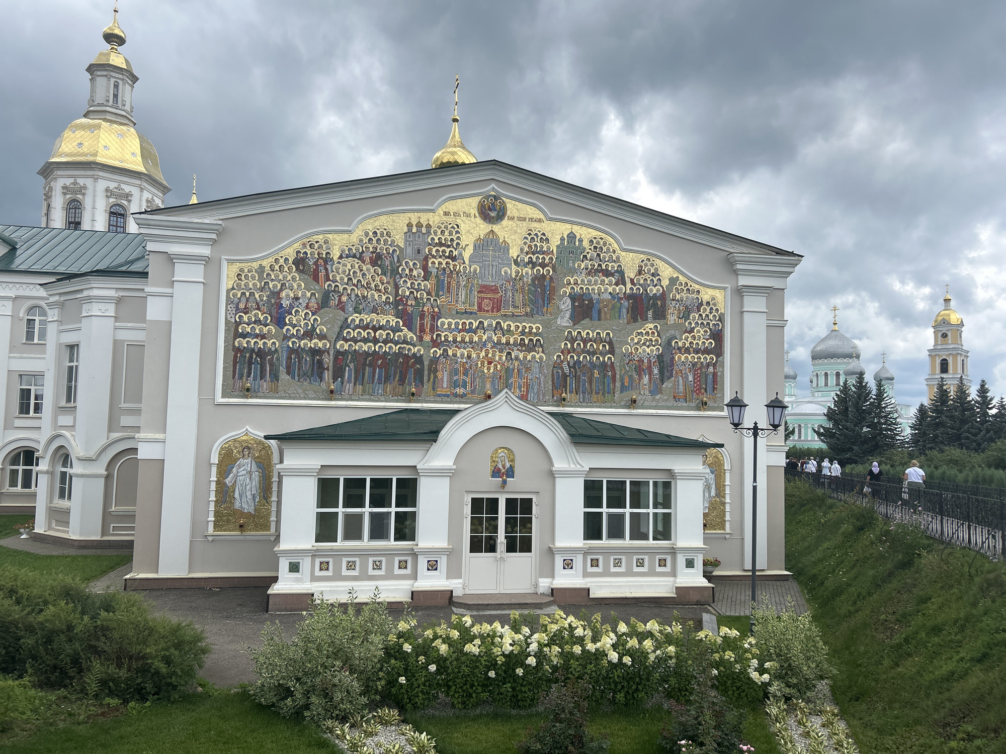 In the city of Diveyevo, Russia, a wide shot captures the exterior of a religious building, likely a monastery or church, under a cloudy sky. The building is adorned with a large, intricate mosaic depicting numerous figures in halos, arranged in hierarchical rows. Above the mosaic, a golden dome with a cross punctuates the sky. To the left of the main building, a tall, multi-tiered bell tower with a golden dome rises prominently. In the middle ground, a pathway curves along a grassy embankment, with several individuals in casual attire walking along it. The foreground features lush green grass and flowering bushes, adding a touch of natural beauty to the scene.