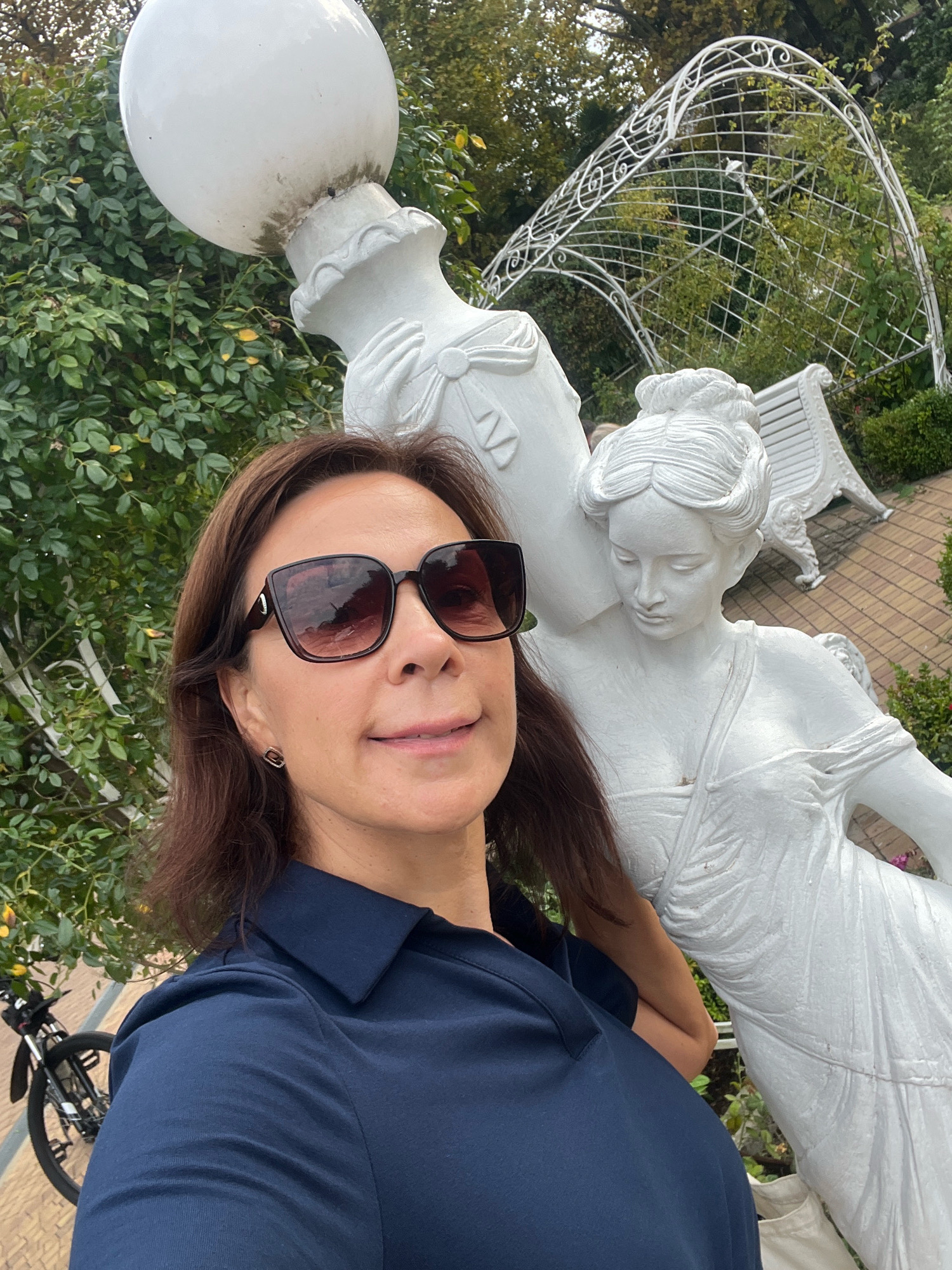 A woman is taking a selfie outdoors, in front of a white sculpture of a woman. The sculpture is in a garden setting with lush green foliage and a white trellis archway in the background. The woman is wearing sunglasses and a navy blue collared shirt. A bicycle is visible on the left side of the image. The overall setting suggests a park or garden area.