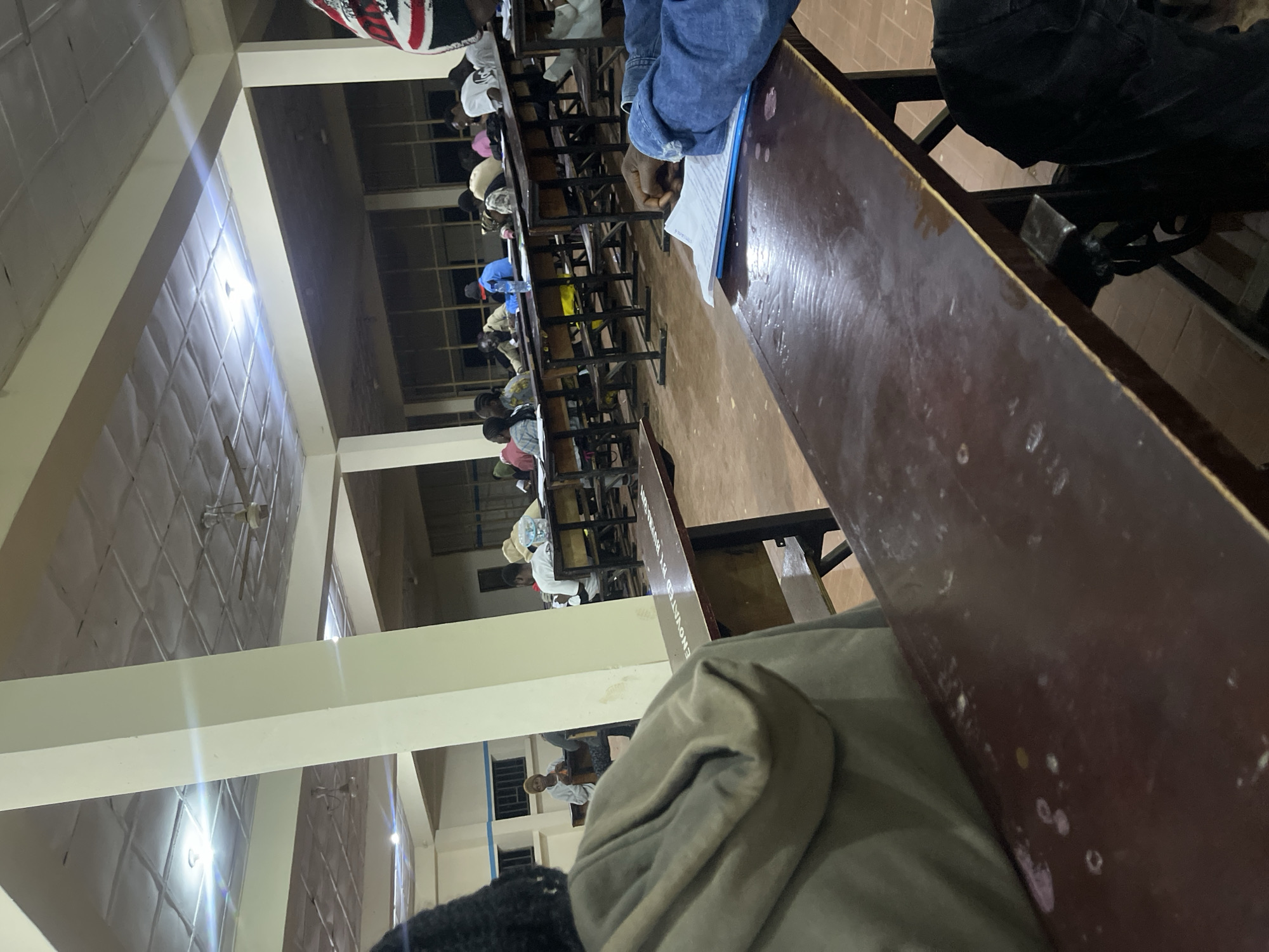 This image depicts an indoor scene, likely a lecture hall, populated with a number of students seated at wooden desks arranged in rows. The environment appears to be dimly lit, with overhead fluorescent lighting and ceiling fans visible. The students are engaged in what appears to be a study session or lecture. The overall aesthetic suggests a university or educational institution in a developing country. The presence of specific architectural elements like the tiled ceiling and concrete pillars, along with the style of the desks, could be characteristic of educational facilities in Nigeria. Without more information, pinpointing the exact location in Rafin Par, Nigeria is not possible, but the scene is consistent with a typical classroom setting within the country.