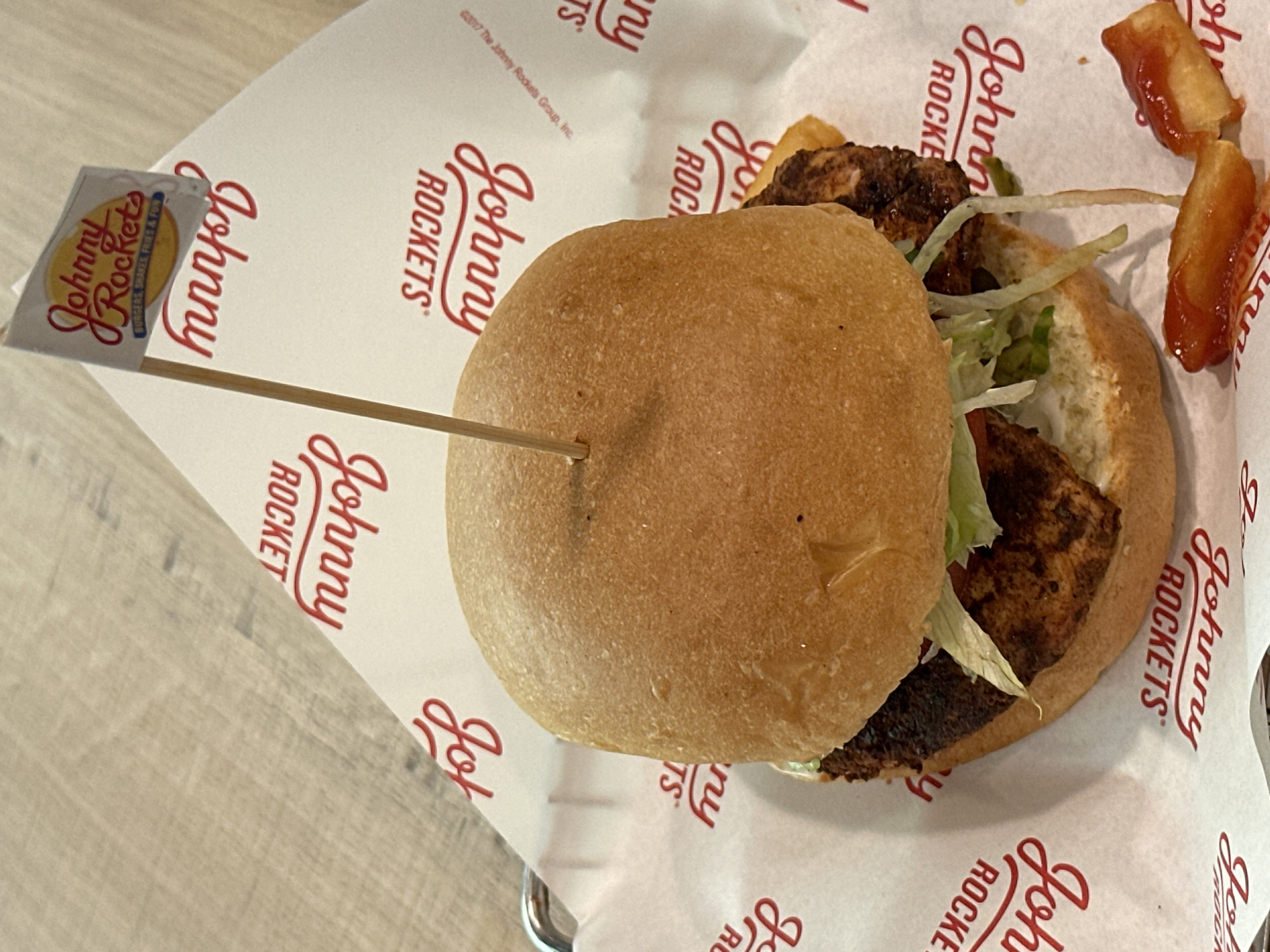 The image shows a Johnny Rockets hamburger. The burger is on a paper liner with the Johnny Rockets logo. The burger is held together with a toothpick that has a Johnny Rockets flag. There are also some fries with ketchup. The location cannot be determined from the image.
