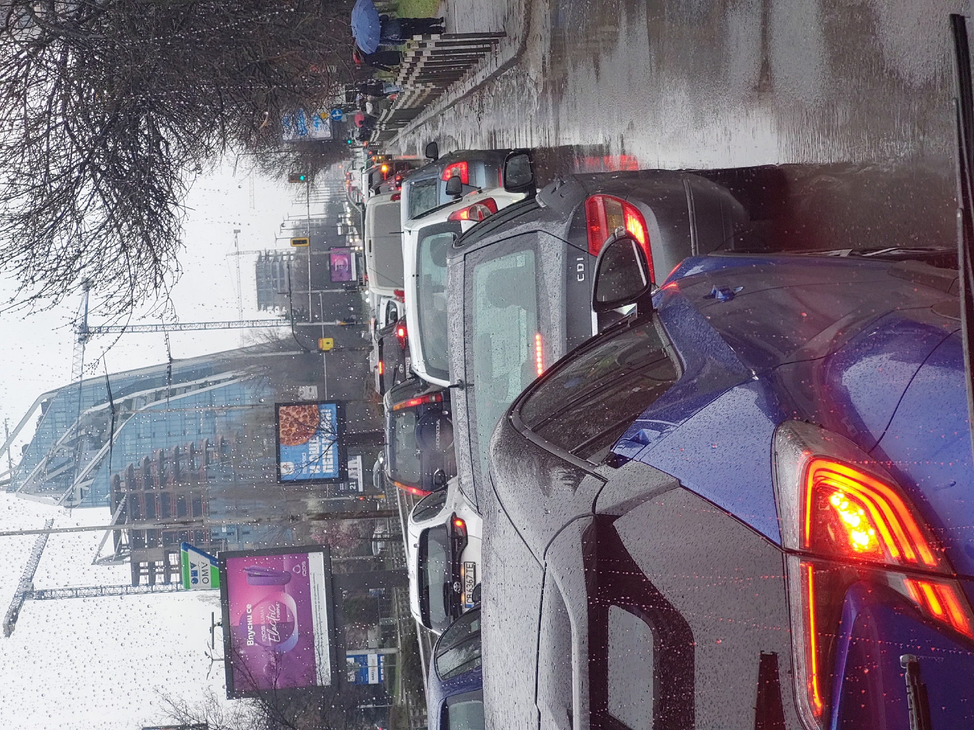 The image shows a traffic jam on a rainy day. The location appears to be in a city, evidenced by the presence of tall buildings, including one under construction with a crane. Billboards advertise products such as pizza and a cigarette alternative. Pedestrians can be seen walking on the sidewalk under umbrellas. The cars are densely packed. The car closest to the camera is blue with its taillight illuminated. The license plates of other cars are partially visible. The environment is wet, as indicated by the rain droplets on the windshield and the wet road surface.