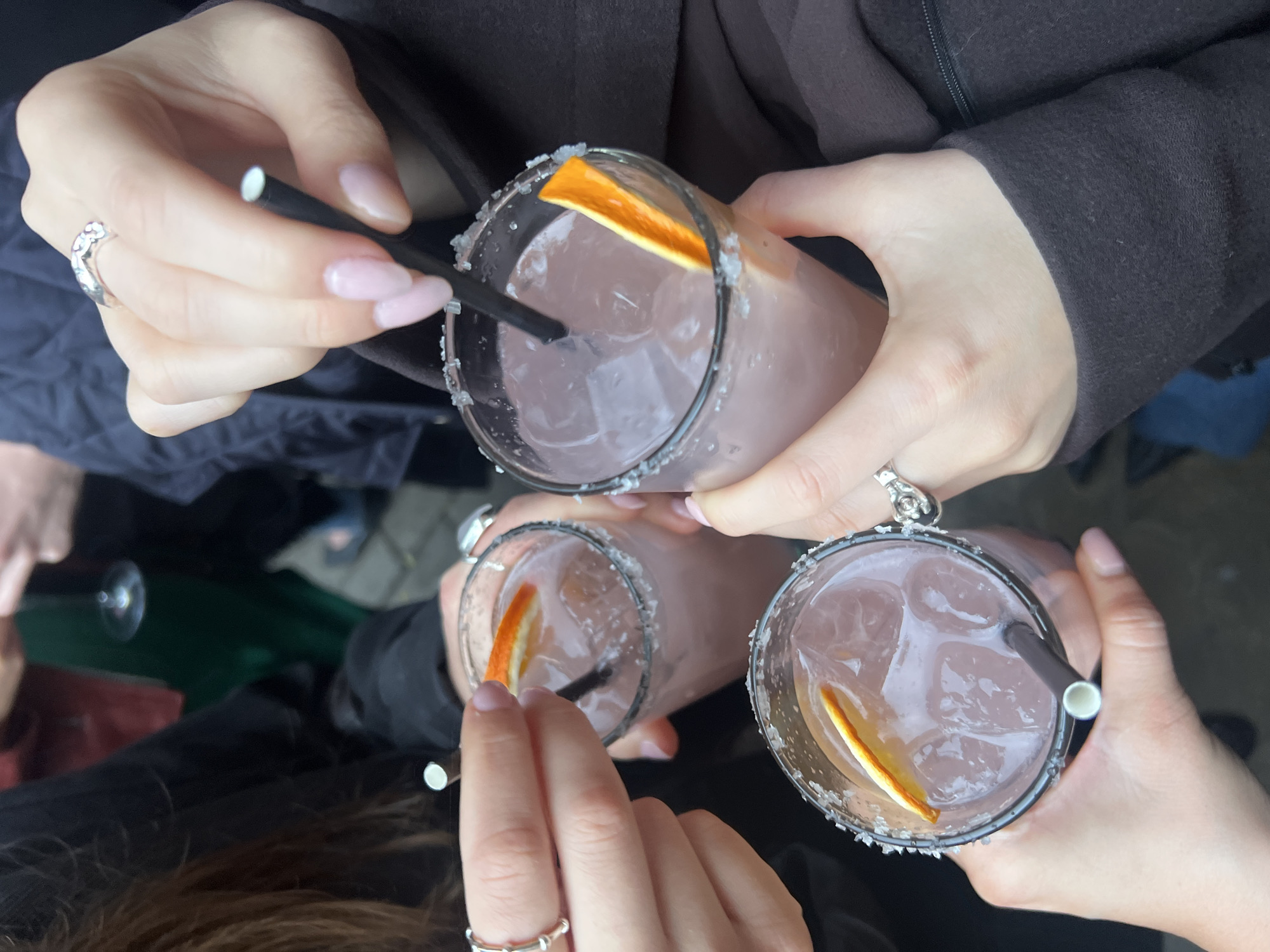 Three people hold glasses with a pink beverage, ice, a black straw, and an orange wedge. The rims of the glasses are covered in white granules. The people are wearing rings on their fingers. They appear to be outdoors. The location is not discernible from the image.