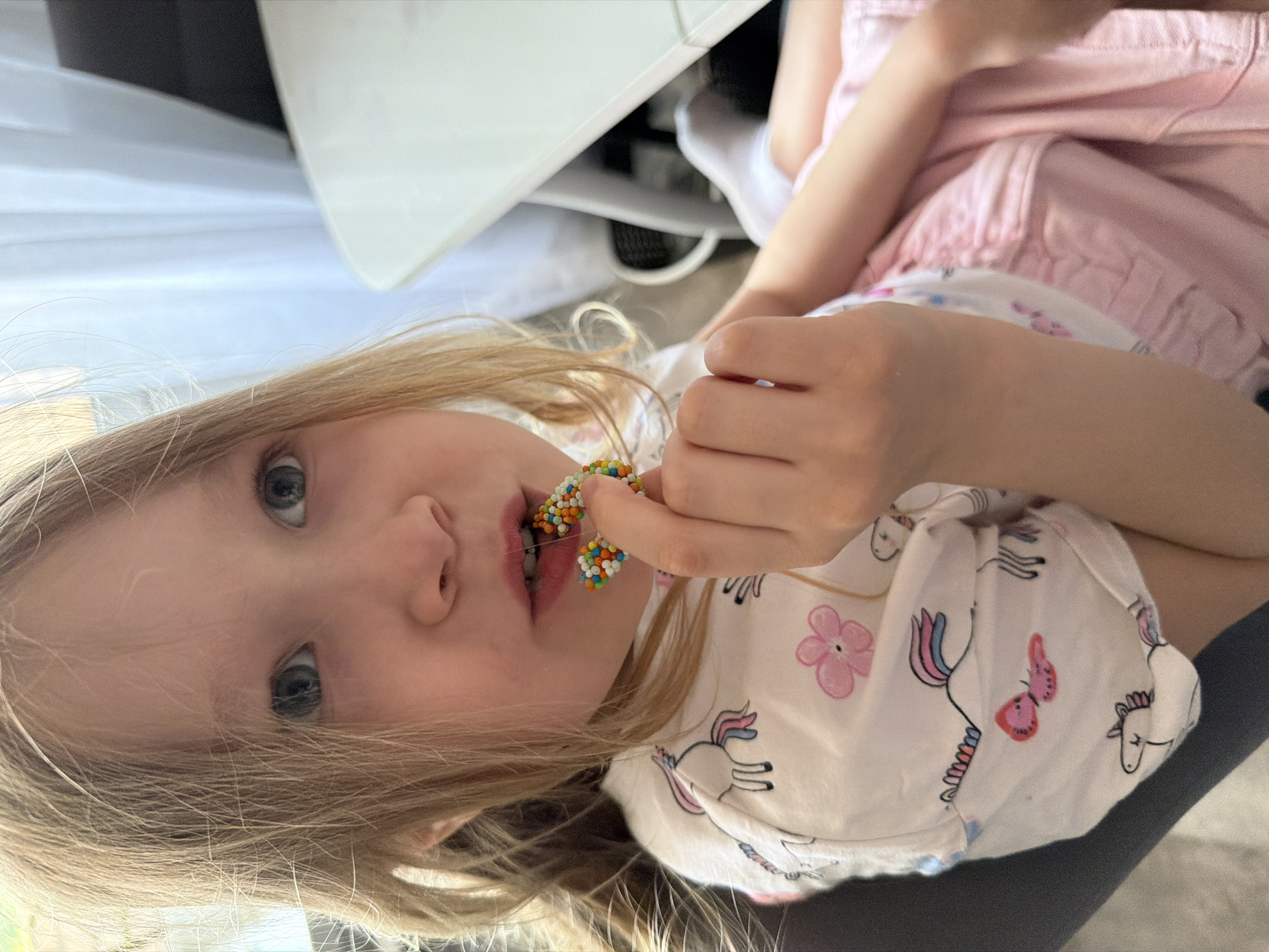 A young girl is seated, holding a small beaded object near her mouth. She has blond hair and blue eyes, and is wearing a white shirt with unicorn and flower patterns, and pink shorts. The background includes a white piece of furniture and curtains. The location is not discernible from the image.
