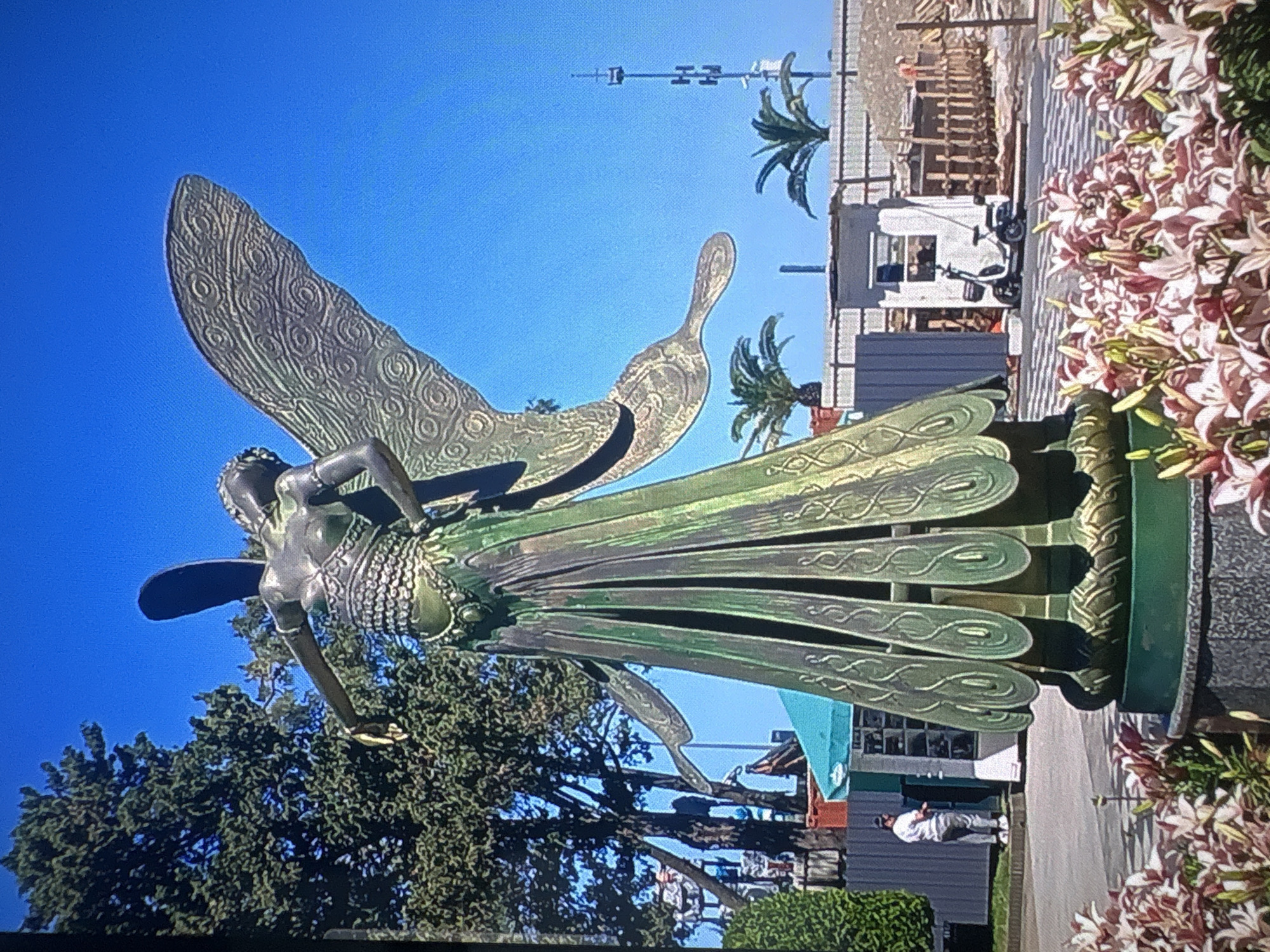 The image captures a large bronze statue of a fairy with outstretched wings, adorned in an ornate dress. The statue is centrally positioned in a park-like setting, with a vibrant blue sky serving as the backdrop. In the foreground, a cluster of lily flowers adds a splash of color. Behind the statue, trees, a turquoise-colored awning, and a tall communications tower are visible. Further in the background, there appears to be construction work with wooden fencing and a small white scooter. A lone individual in casual attire is standing near the awning. This statue is located in Batumi, Georgia, on the waterfront promenade.