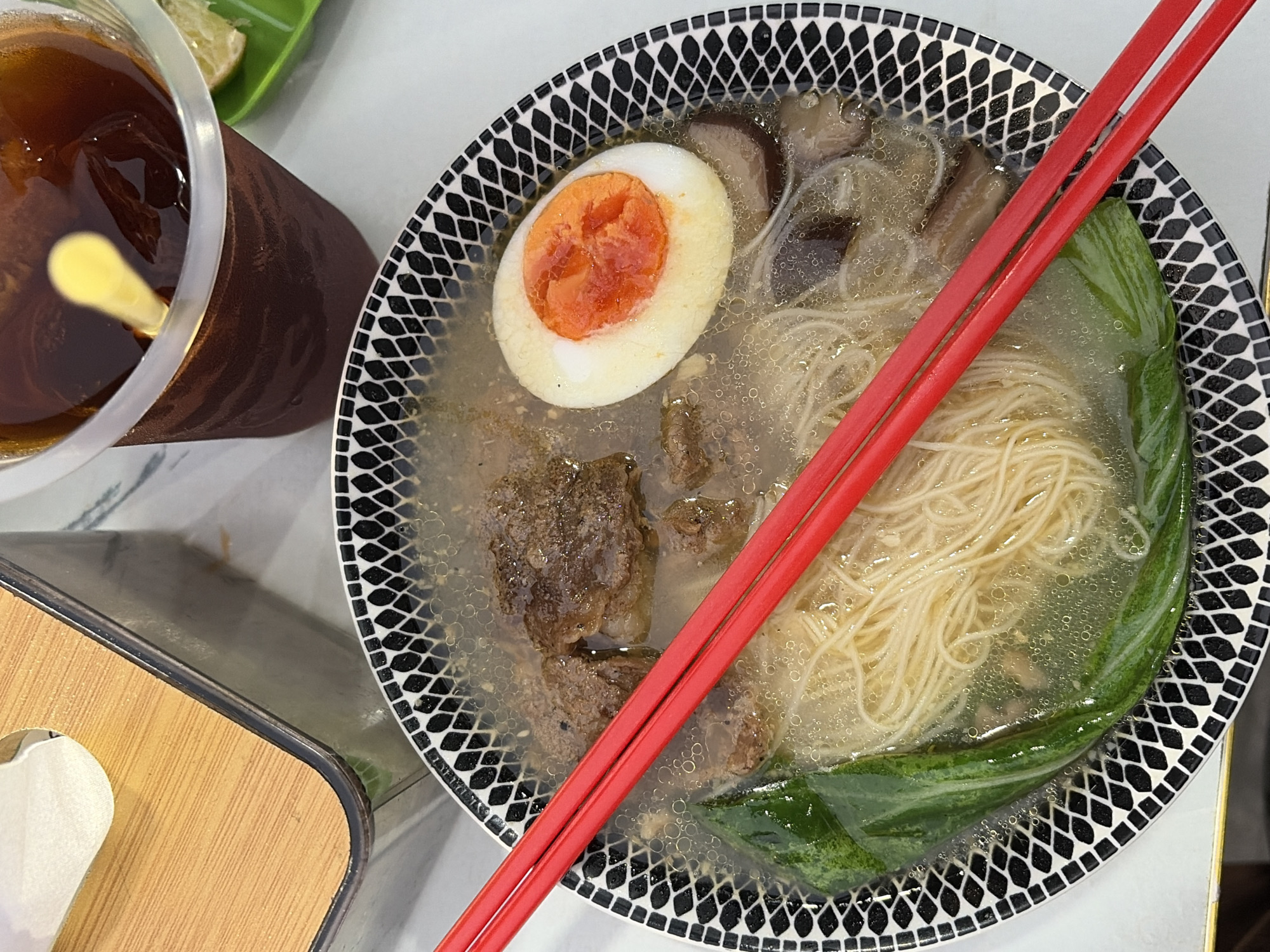 This image captures a close-up overhead view of a bowl of ramen and a drink, likely taken at a restaurant or eatery. The ramen is served in a black and white patterned bowl and contains noodles, chunks of meat, a halved hard-boiled egg with a vibrant orange yolk, and green leafy vegetables. Red chopsticks are placed diagonally across the bowl. In the background, a clear plastic cup filled with a dark liquid and ice, with a yellow straw, is visible. To the left, part of a wooden tissue box with a metal rim is also present. The scene suggests a casual dining experience.
Based on the provided information that the image is from Semarang, Indonesia, this scene could be from a local restaurant specializing in Asian cuisine, potentially offering ramen or noodle dishes. The presence of a tissue box is common in dining establishments, and the overall presentation points to a typical meal setting within the city.