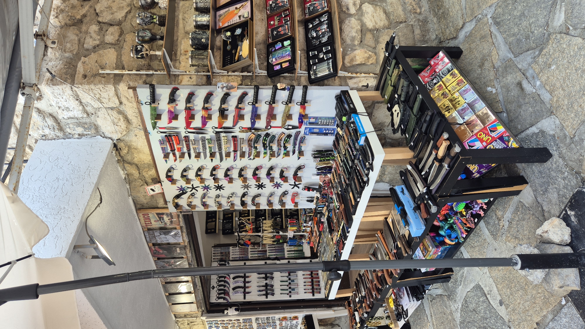 The image shows an outdoor market stall in Nesebar, Bulgaria, selling knives and other accessories. The stall is set up against a stone building, with a white awning overhead. The merchandise is displayed on white tables and black shelves. On the tables, there are rows of knives of various sizes and designs, some with colorful handles. There are also some novelty items like toy dinosaurs. The shelves display more knives, as well as some packaged items like playing cards and liquor bottles. In the background, there are more shelves with decorative items and packaged goods. A black umbrella pole stands in the foreground, partially obscuring the view of the stall. The ground is paved with stone tiles.