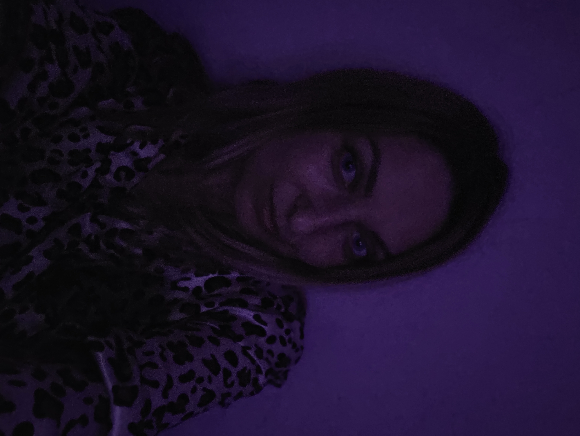 A single female individual is depicted in an indoor setting, positioned centrally within the frame against a plain wall. Her head and upper torso are visible. She has light-colored eyes and long hair that appears dark, partially obscuring the left side of her face due to shadow. Her expression is neutral. She is wearing a garment featuring a dark, possibly black, leopard print pattern on a lighter, dark purple background, visible across her right shoulder and chest. The scene is illuminated by dim, purple-hued lighting, casting significant shadows, particularly on the left side of her face and body. The background consists of a uniformly textured wall, saturated in a dark purple color due to the ambient light. No other subjects, objects, or specific environmental features are discernible.