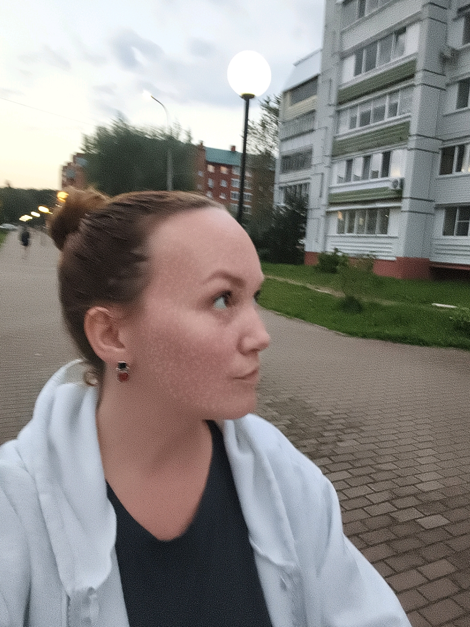 A medium shot captures a woman from the chest up, with her head turned to the right, in an outdoor urban setting. She has her hair pulled up in a bun and is wearing a white hooded jacket over a dark top and an earring in her left ear. The background shows a modern apartment building with multiple windows, a paved walkway, and green grass. The sky is overcast.