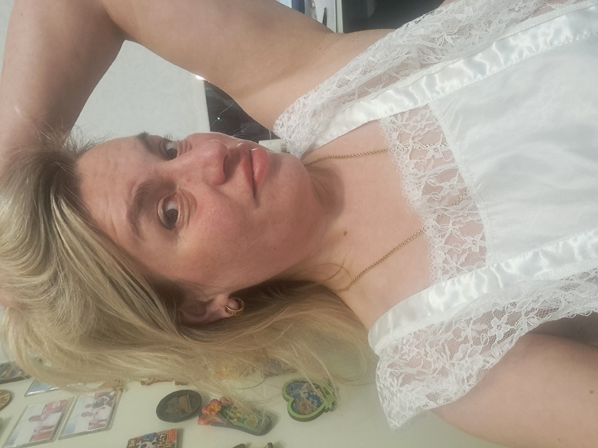 A woman with blonde hair is lying down wearing a white, lace-trimmed garment. Her head is tilted, and her arm is raised near her head. A gold necklace is visible on her neck. The background appears to be a wall with magnets attached. There is no information to suggest that this image was taken in Elektrostal, Russia.