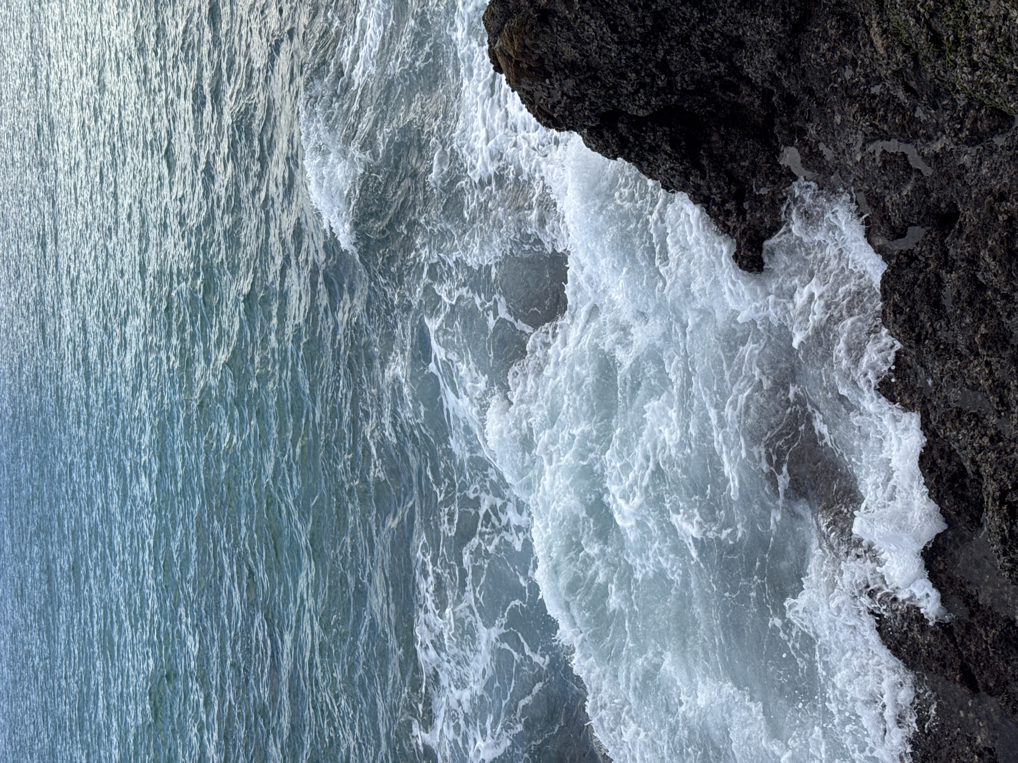 The image shows a body of water with waves crashing against dark, rocky shore in the foreground. The water is a deep blue-green, with white foam created by the crashing waves. The rocks are rough and textured, with some appearing to be covered in algae or seaweed. The sky is not visible in the image, but the lighting suggests it is daytime. The image is a close-up shot, focusing on the interaction between the water and the rocks. There are no people or man-made objects visible in the image. The location is not specified in the city of Choeng Thale, Thailand.