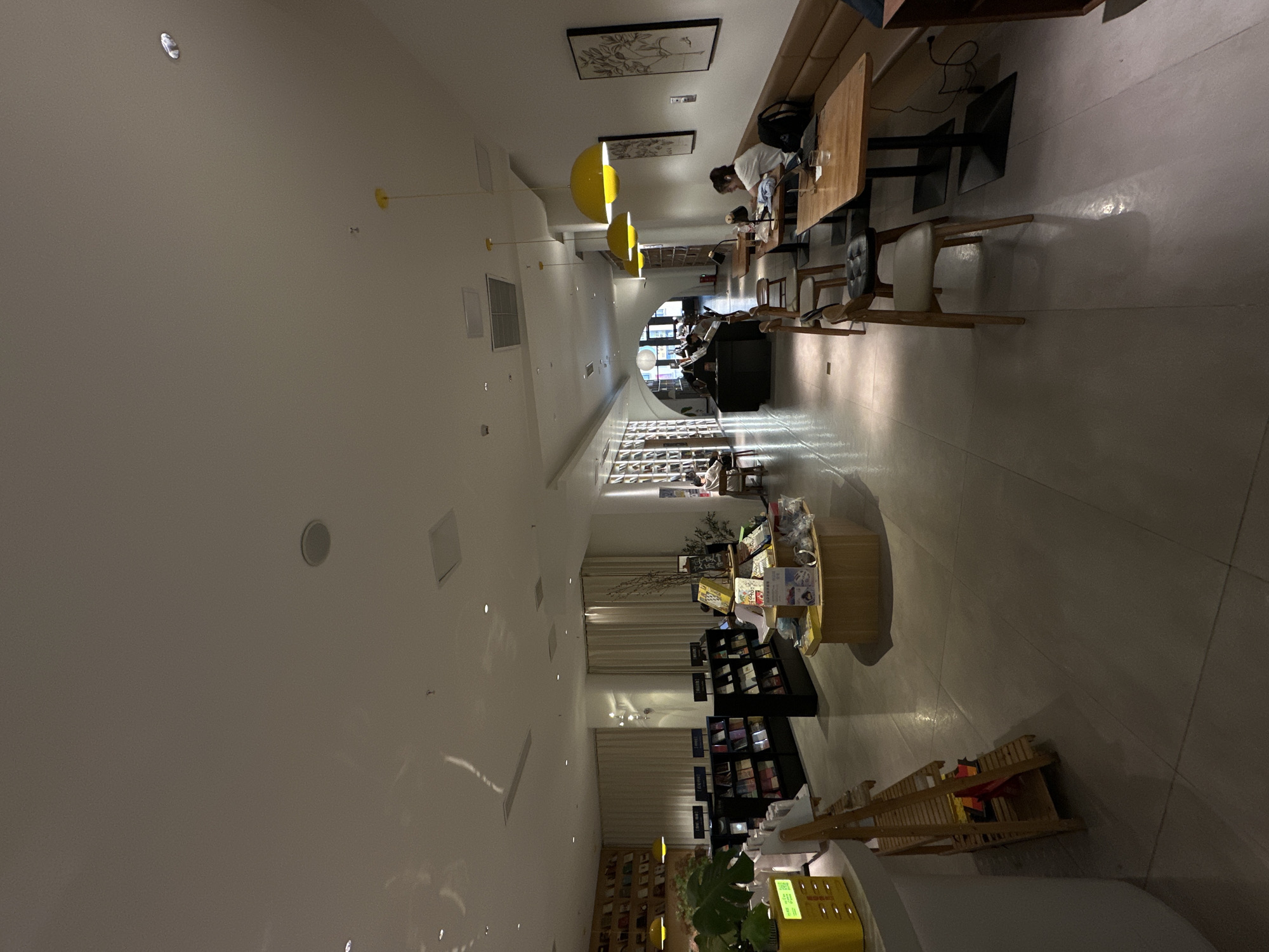 This image depicts the interior of a modern bookstore and cafe, likely located in the Chongqing High-tech Zone, China. The space is brightly lit with a combination of overhead and accent lighting, casting reflections on the polished concrete floor. In the foreground, a wooden shelving unit with some items on display stands on the left. Further back, a circular display stand showcases books. Rows of bookshelves line the walls, filled with books. Several tables and chairs are arranged throughout the space, with some patrons visible. On the right side of the image, a long bench seating area runs along the wall, accompanied by wooden tables where individuals are seated, appearing to be reading or working on laptops. Yellow pendant lights hang above some of the seating areas. The overall atmosphere suggests a quiet and contemplative environment conducive to reading and studying.