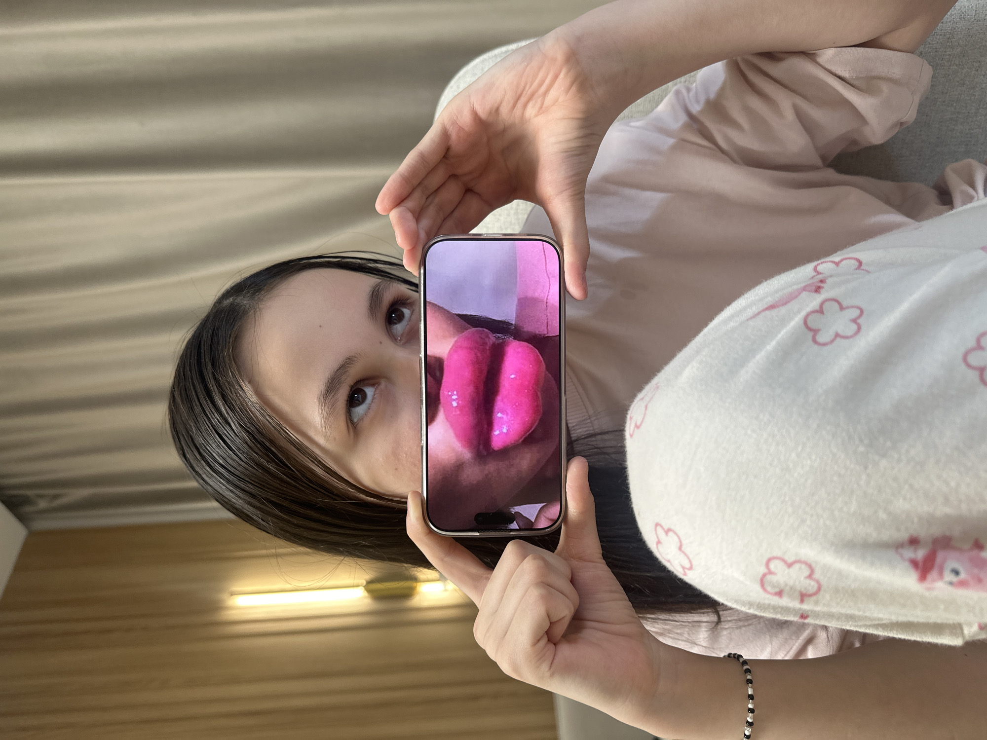 The image, taken in Votkinsk, Russia, is a portrait-oriented shot, rotated 90 degrees counter-clockwise, showing a young person lying down in a relaxed indoor setting. The individual, with long dark hair, is visible from the chest up, wearing a light pink long-sleeved top and white pajama bottoms adorned with pink flower and cartoon animal (possibly bear) patterns. A black and white beaded bracelet is worn on her right wrist.
She holds a modern smartphone with both hands directly in front of her face. The phone's screen displays a close-up image of two vibrant pink, glossy, rounded objects, resembling either stylized lips or a heart shape. The person's eyes are directed upwards and to the right, looking past the smartphone. The background behind her head features a light beige curtain or textured wall. To the left side of the frame (when upright), a section of wooden paneling and a horizontal light fixture are visible. The overall scene suggests a casual and comfortable atmosphere, likely at home. There is no discernible text visible in the image.
